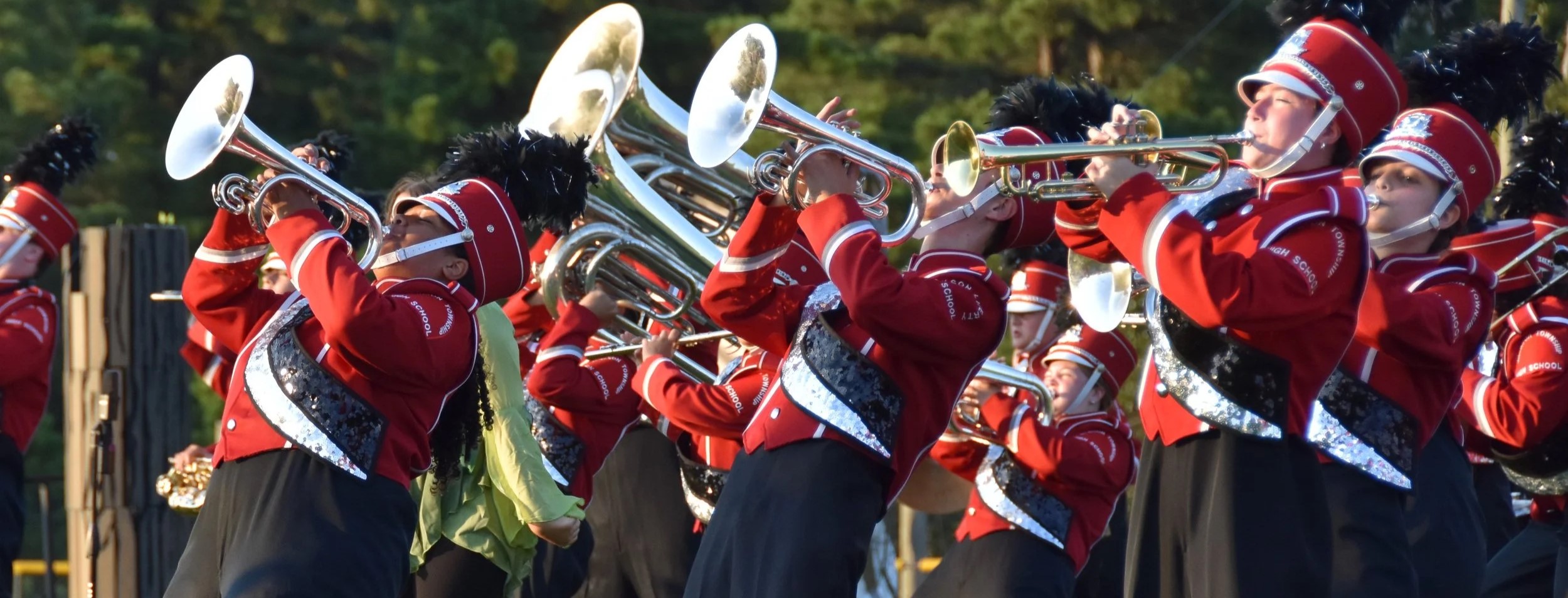 A marching band in bright red and white uniforms performing outdoors, with a forested background.
