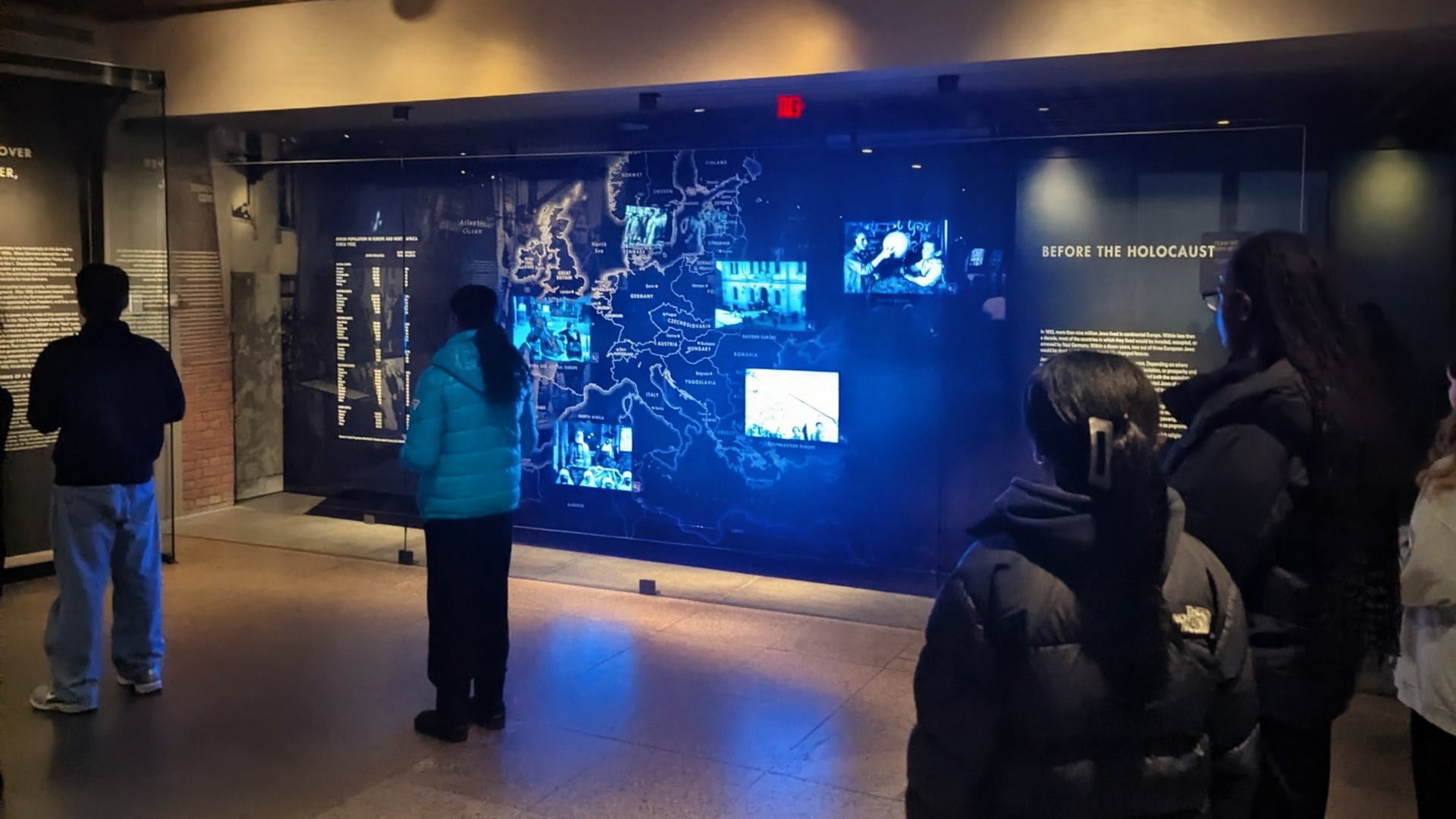 The image depicts a dimly lit museum exhibit space with a large, illuminated display on the wall showcasing various historical artifacts and information. Several people can be seen in the foreground, examining the exhibit.