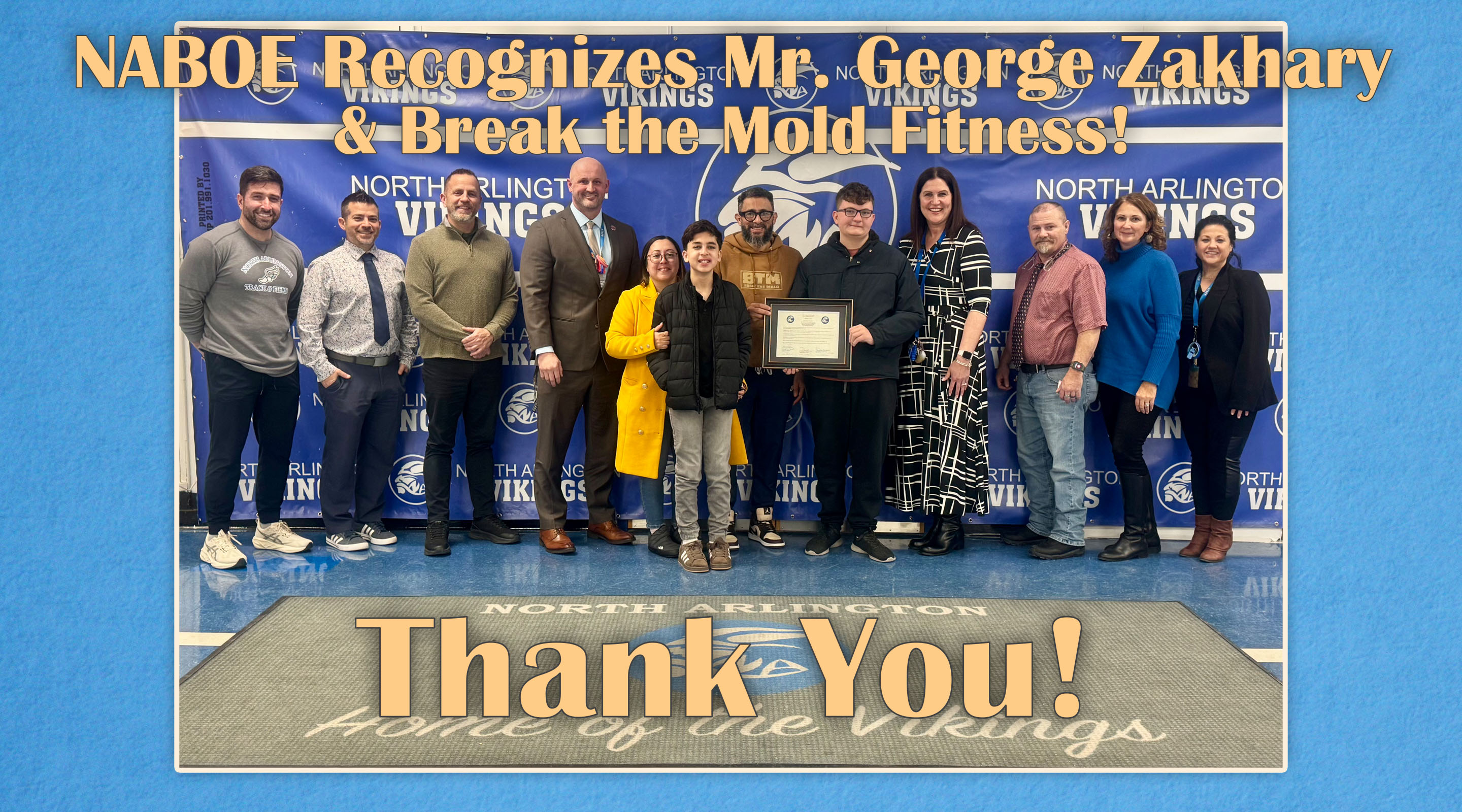 A group of people stand on a gym floor with a "Thank You!" banner in the foreground, while a banner in the background reads "NABOE Recognizes Mr. George Zakhary &amp; Break the Mold Fitness!".