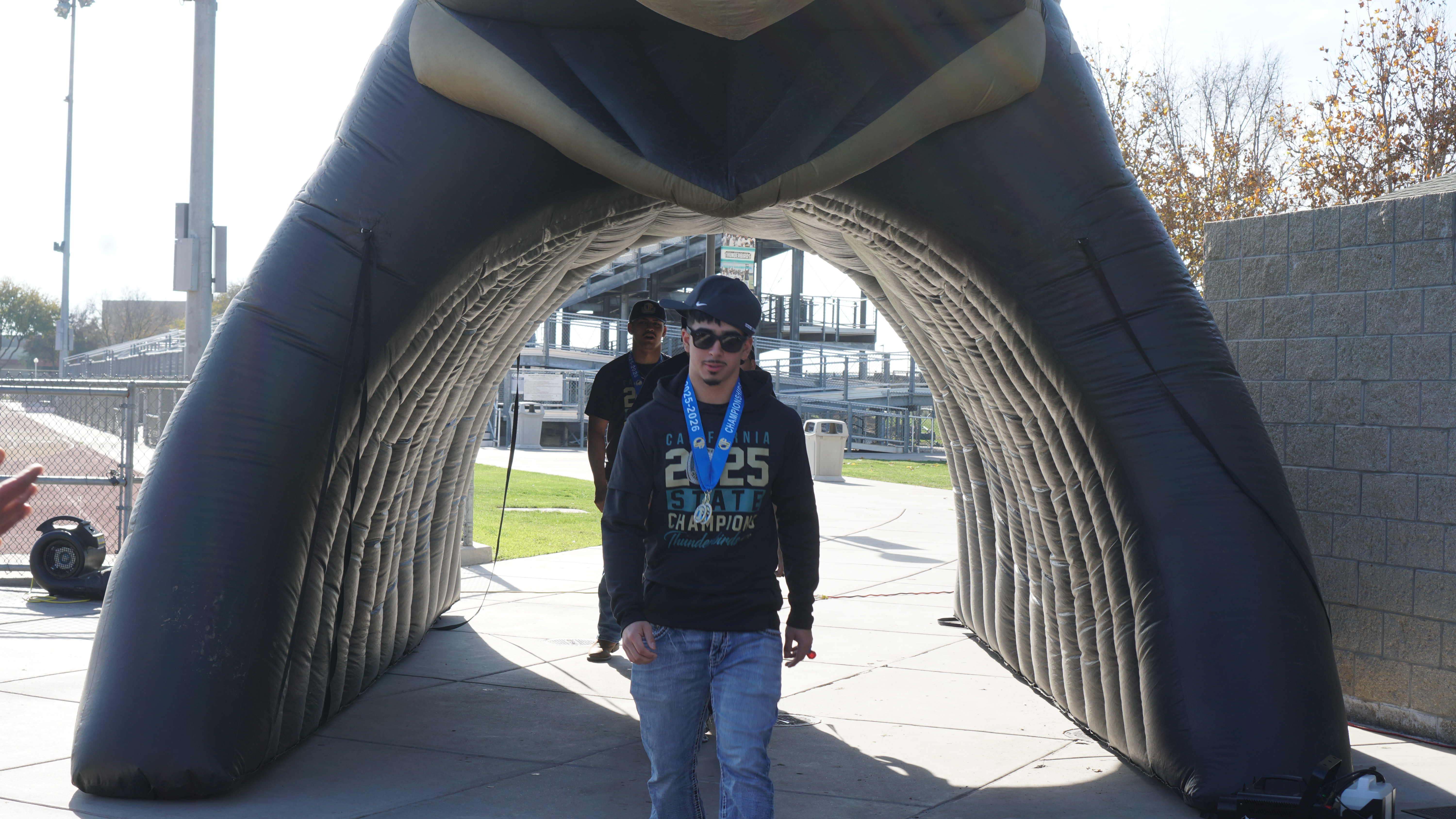 A person wearing a black jacket and jeans stands in an arched entryway, with a grassy area and buildings visible in the background.