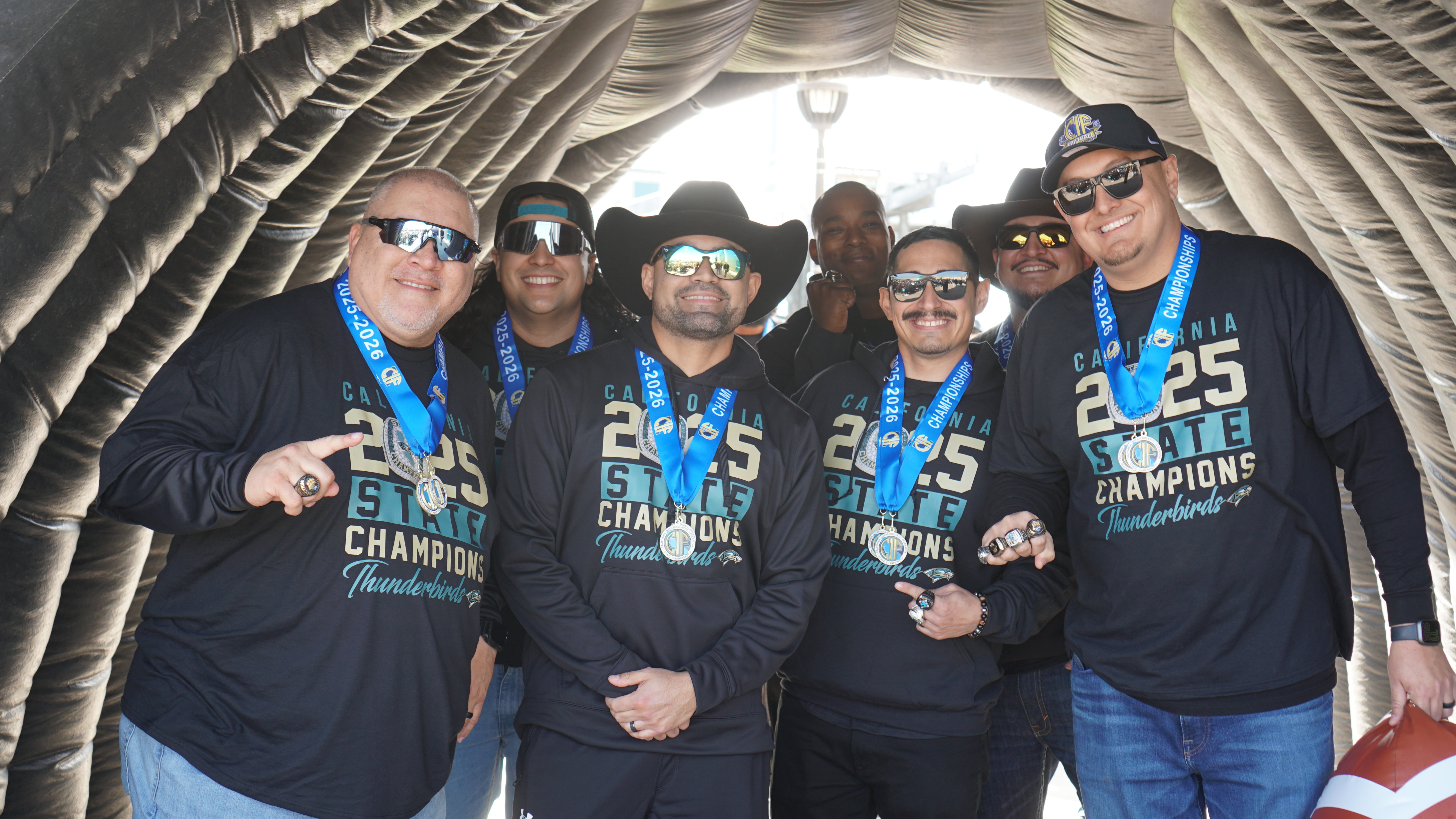 A group of people wearing matching black shirts with "2023 Champions" printed on them, posing together in what appears to be a curtained or draped setting.
