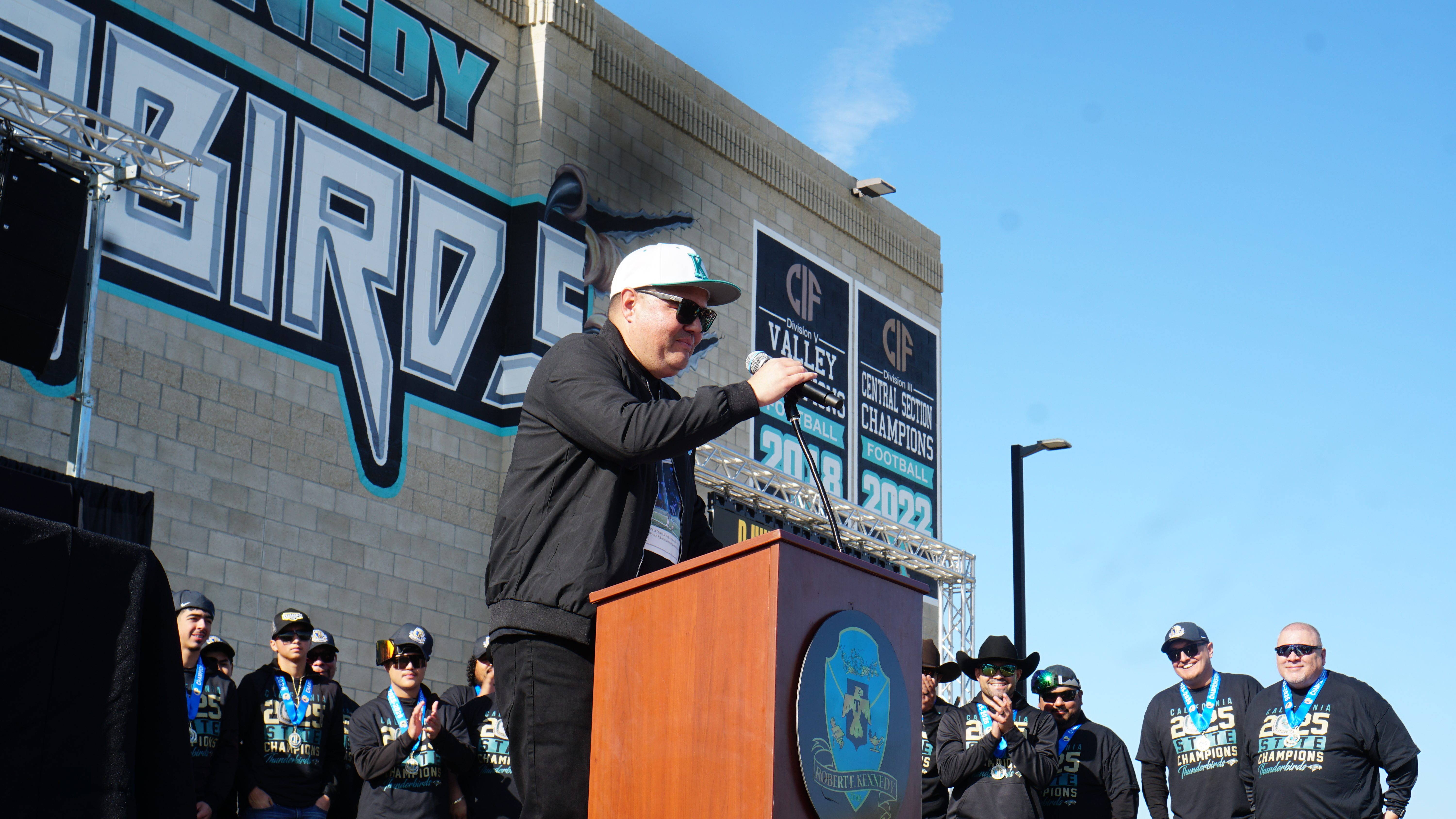 A person in a black jacket and hard hat is standing at a podium in front of a large mural with the word "VICTORY" prominently displayed, surrounded by a crowd of people wearing various sports team apparel.
