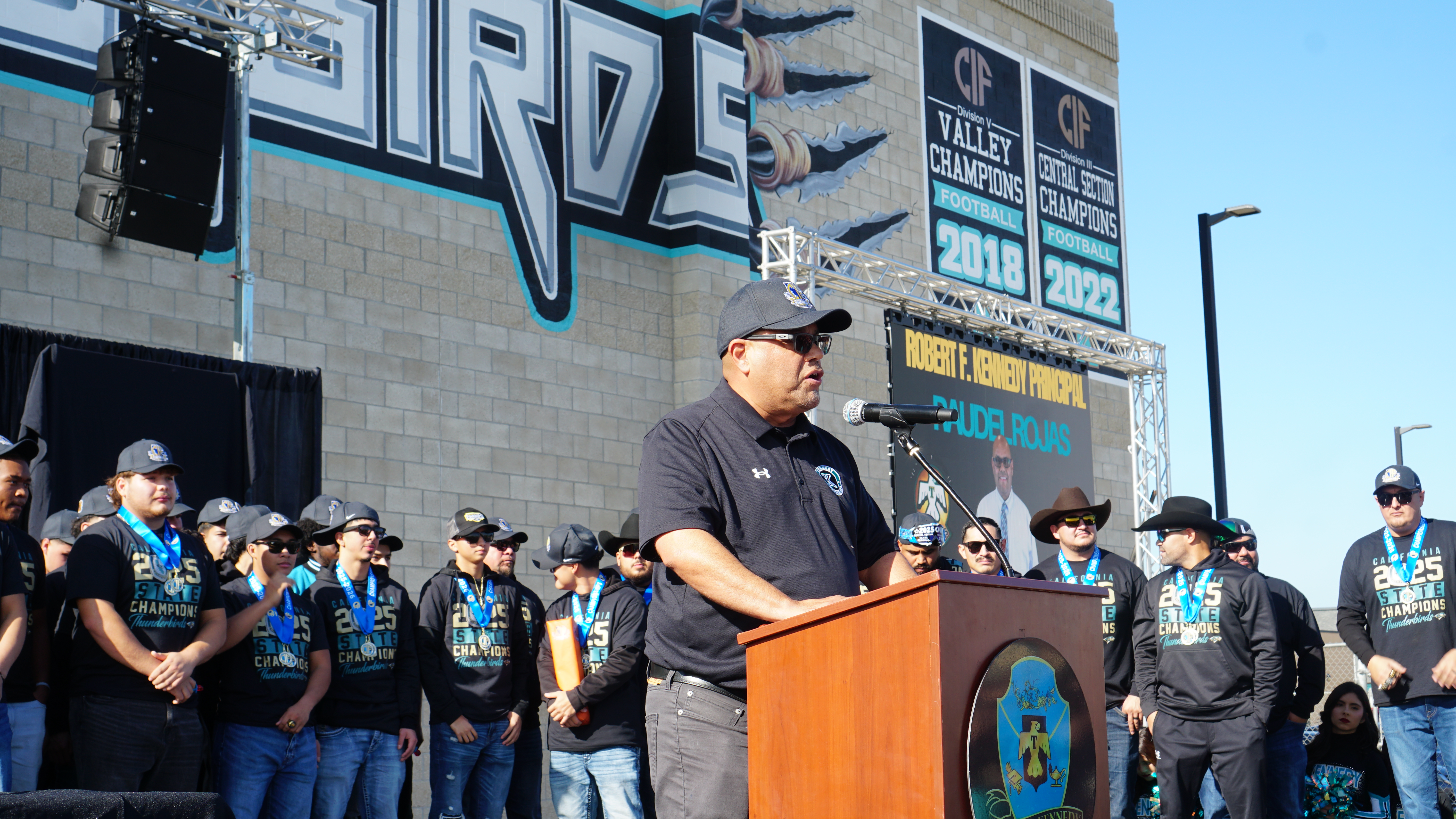 A man stands at a podium in front of a large crowd of people wearing matching uniforms, with a large graphic sign visible in the background.