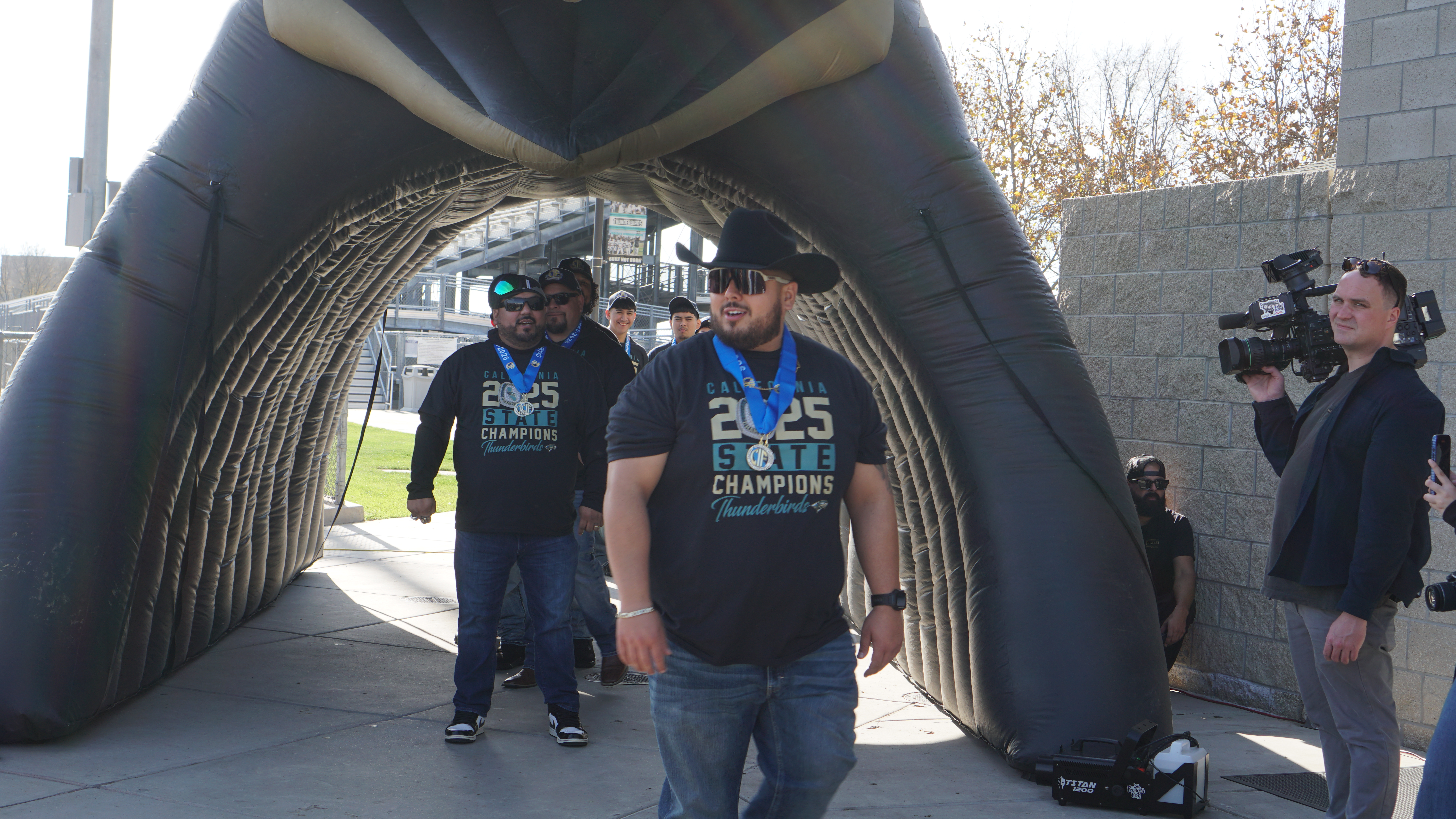 The image shows a group of people walking through an arched structure, with a person in the foreground wearing a t-shirt with the text "Campus Champions" and others in the background.