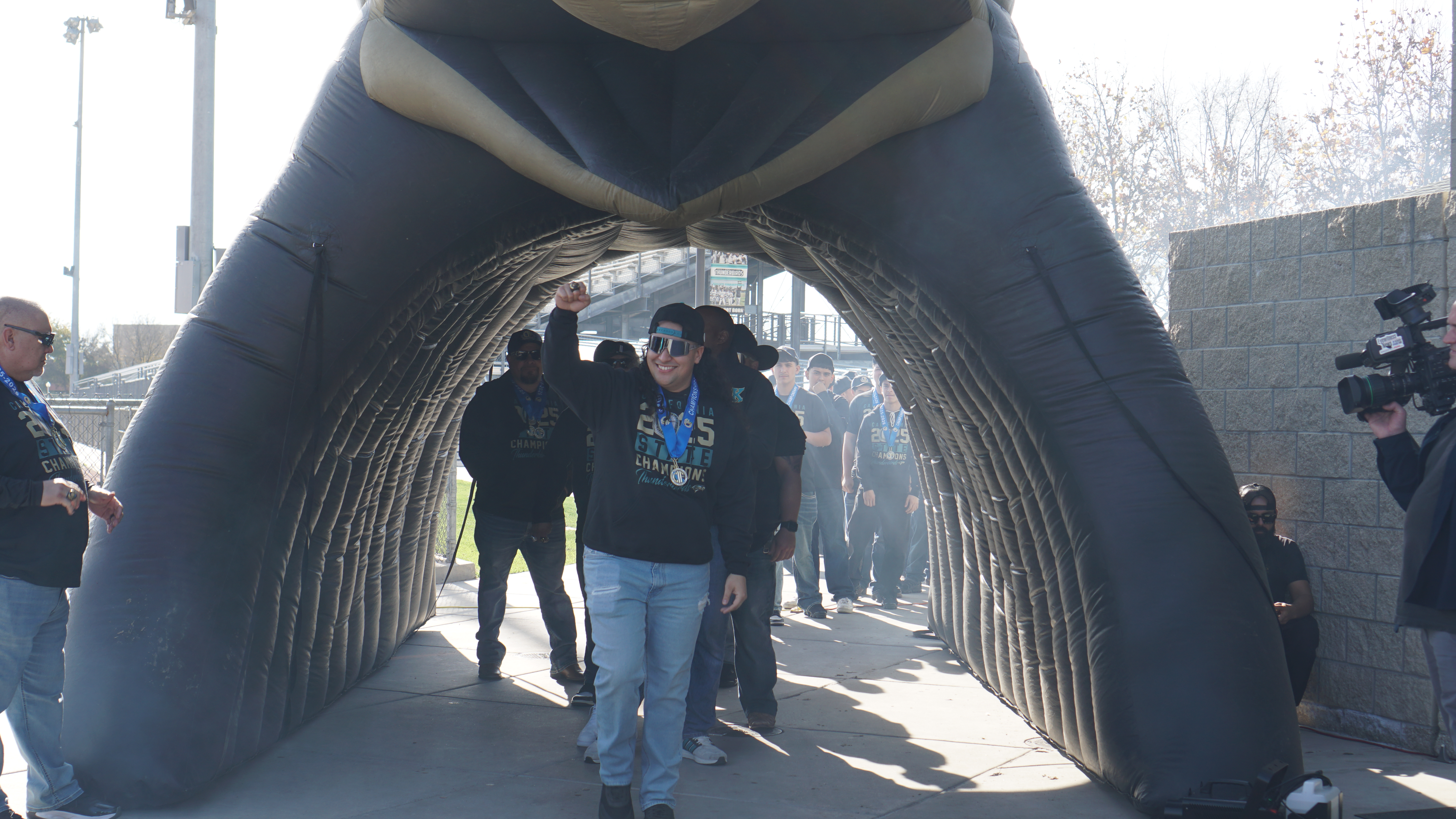 A group of people walking through an arched structure made of metal rods, with a cloudy sky and buildings visible in the background.