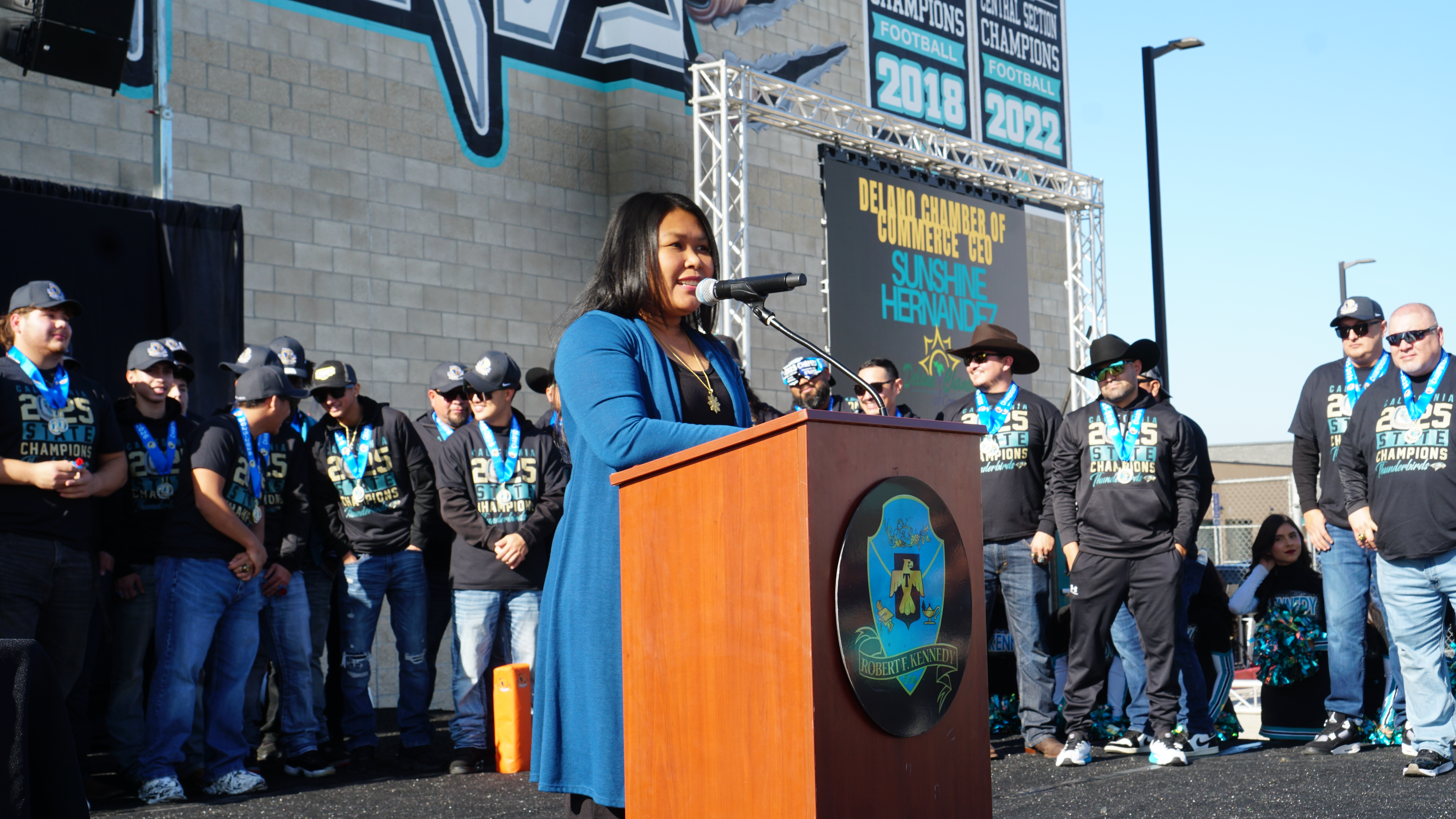 A woman in a blue coat is standing at a podium, speaking to a crowd of people wearing various uniforms and casual attire in the background.