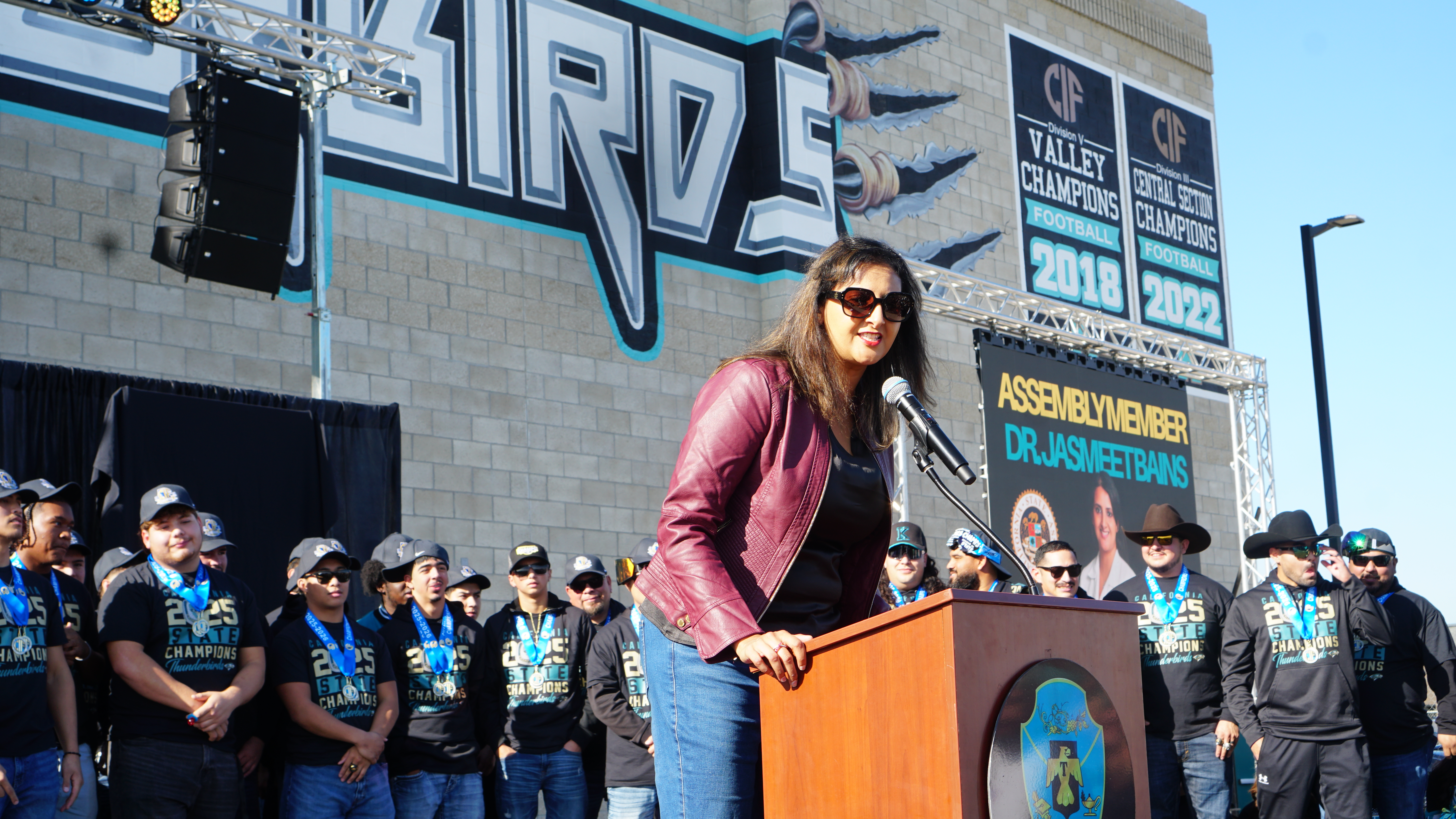 A woman in a pink jacket stands at a podium in front of a large crowd, with a graffiti-covered wall in the background.
