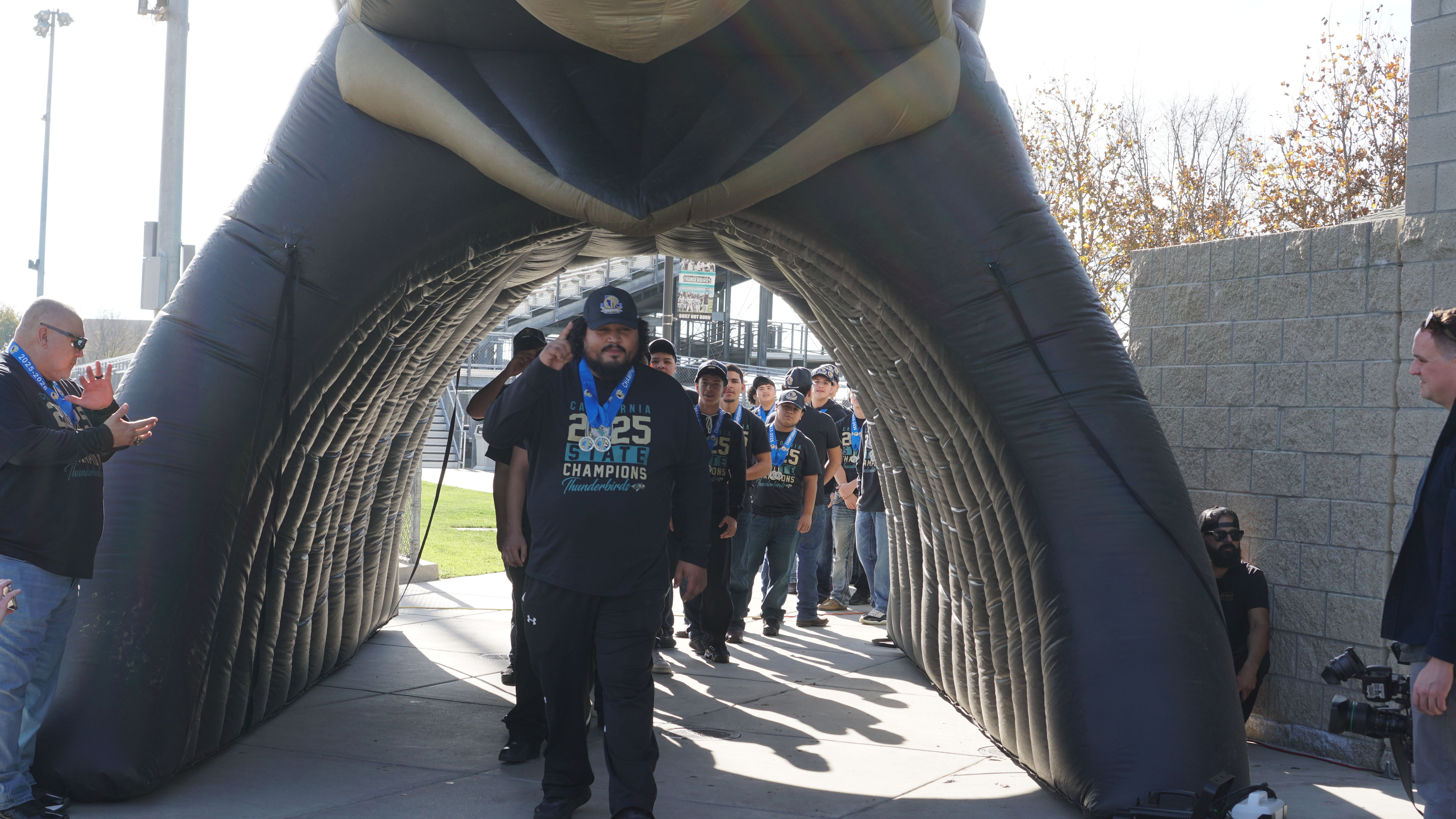 A group of people, some wearing sports jerseys, are walking through an arched entryway with a large, abstract sculpture in the foreground.