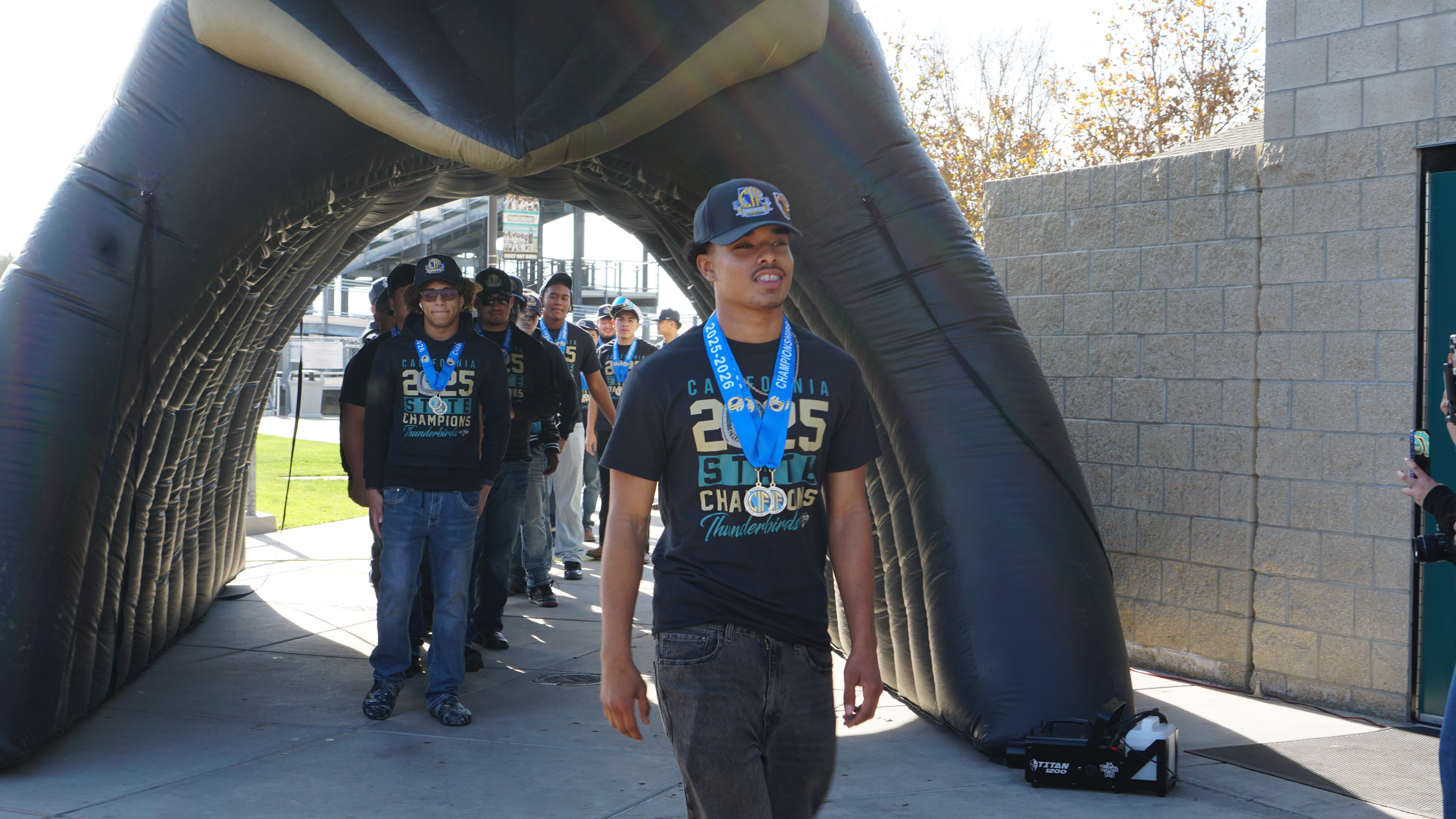 The image shows a group of people walking through an archway, with one person in the foreground wearing a blue jersey and carrying a skateboard.