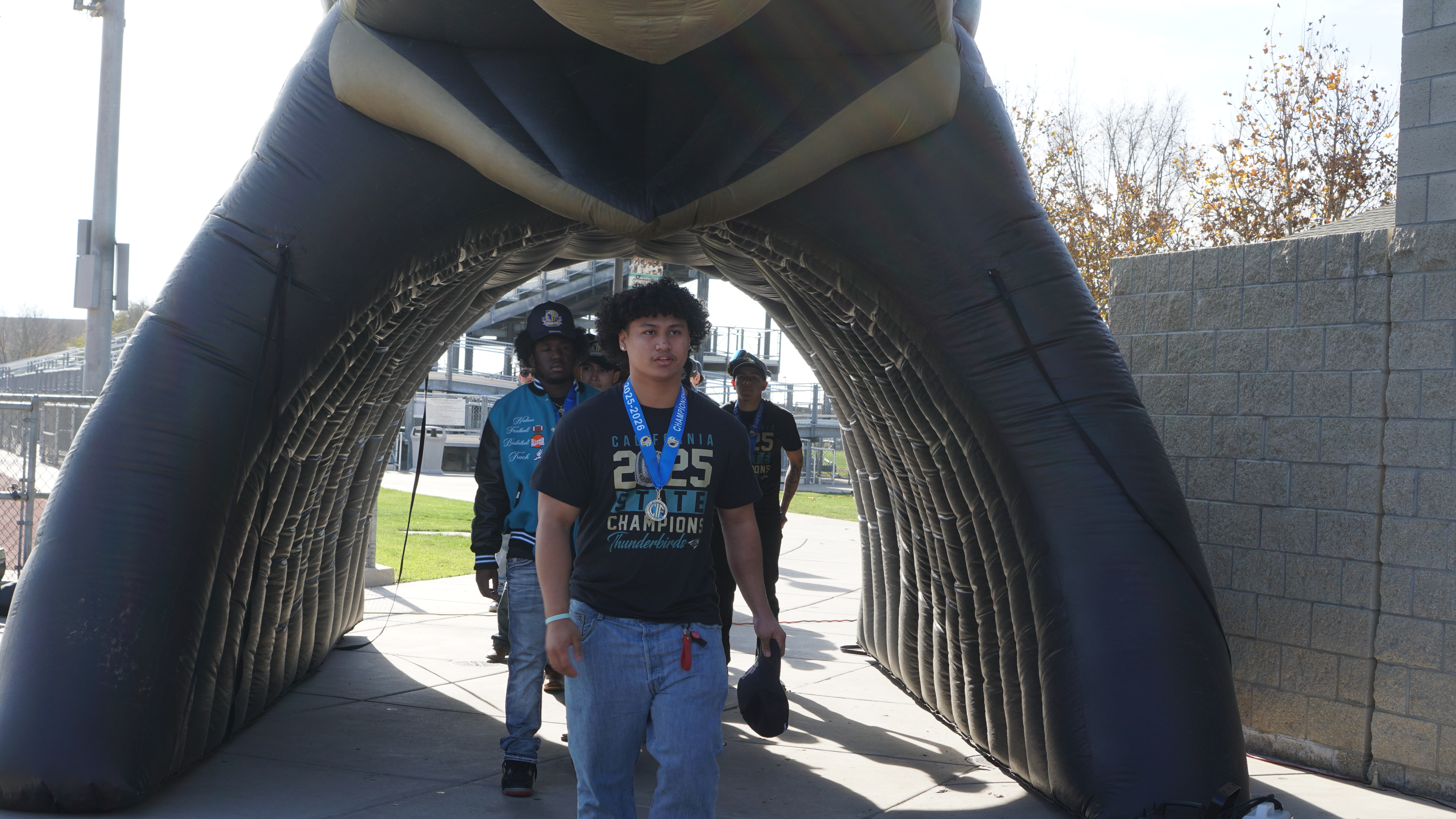 The image shows a young person wearing a blue shirt and jeans standing in an archway-like structure, with a background of trees and buildings visible.