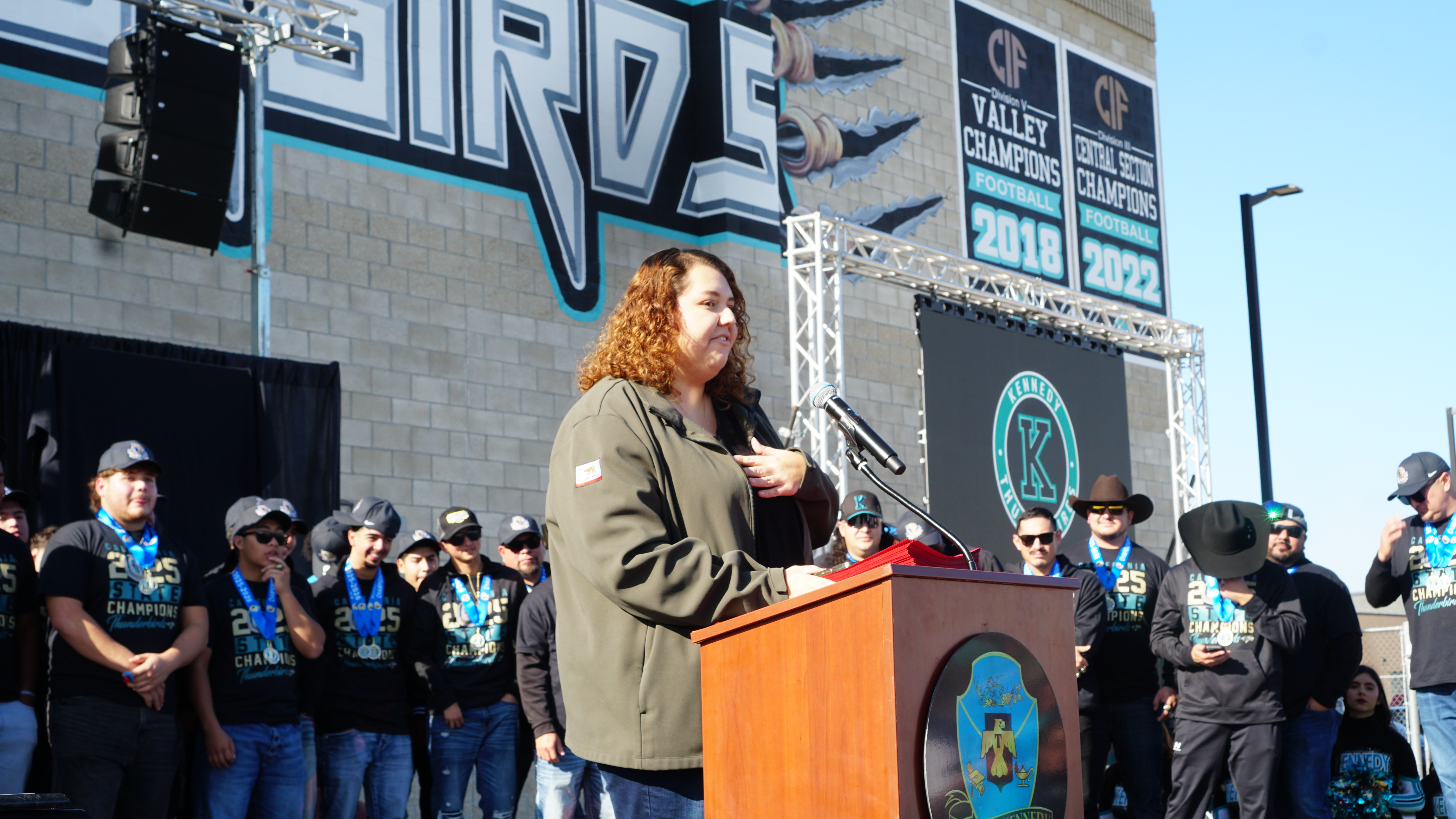 The image shows a person with long, curly red hair standing at a podium in front of a large crowd, with a graffiti-covered wall in the background.