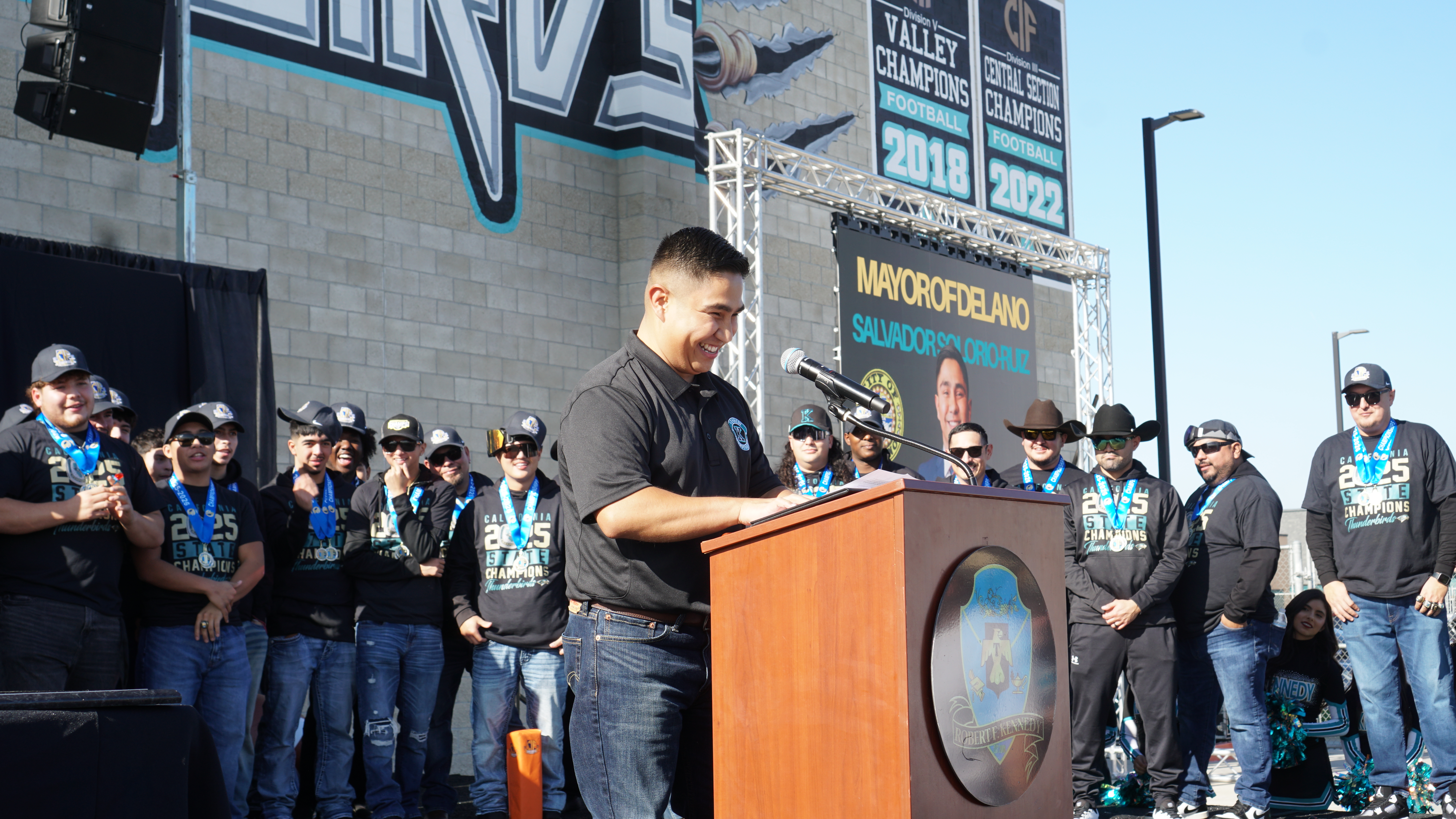 A man is standing at a podium addressing a crowd of people wearing sports jerseys and hats in front of a large building with signage.