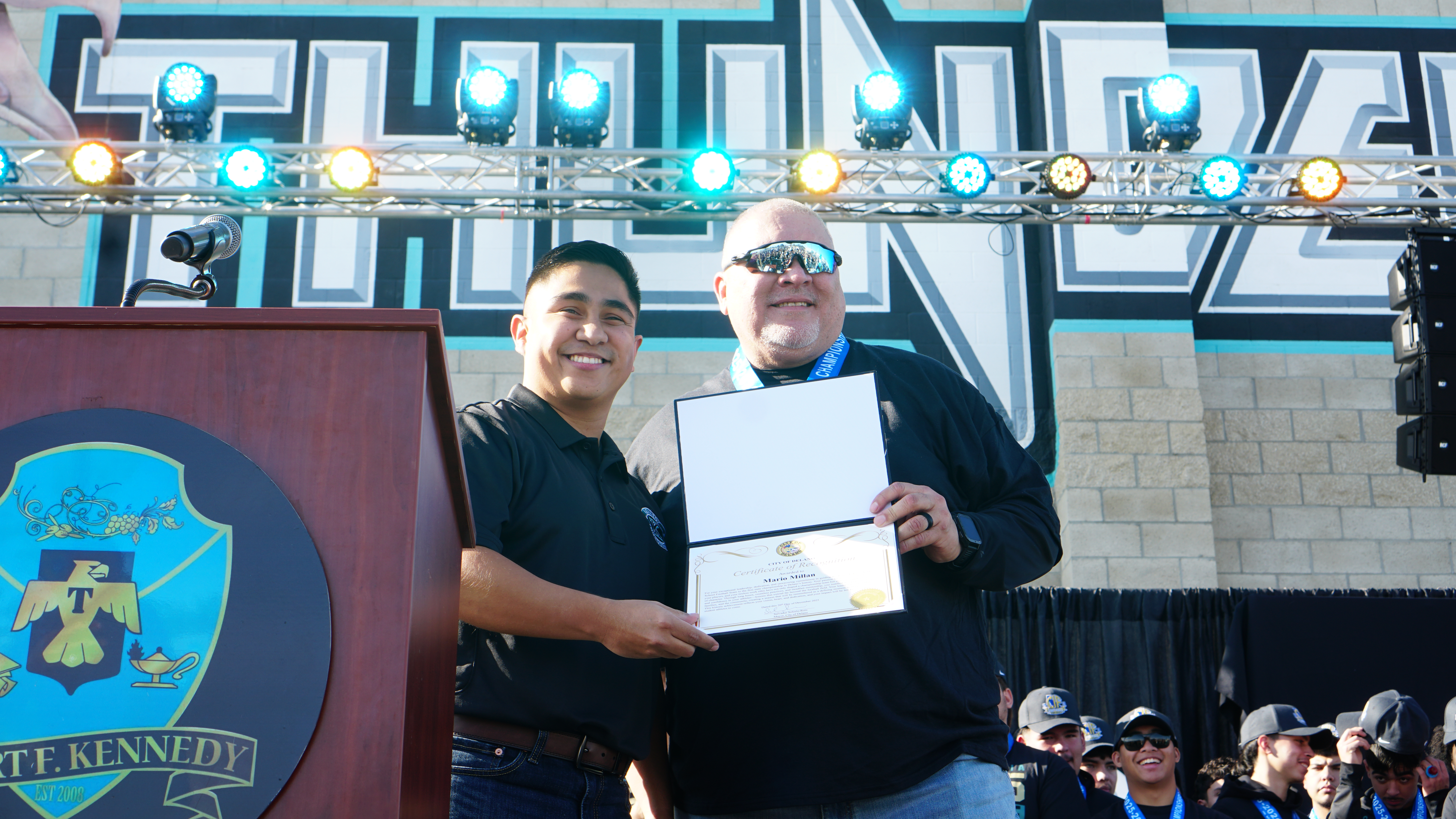 Two men in formal attire are standing in front of a backdrop with a coat of arms and decorative lighting fixtures in the background.