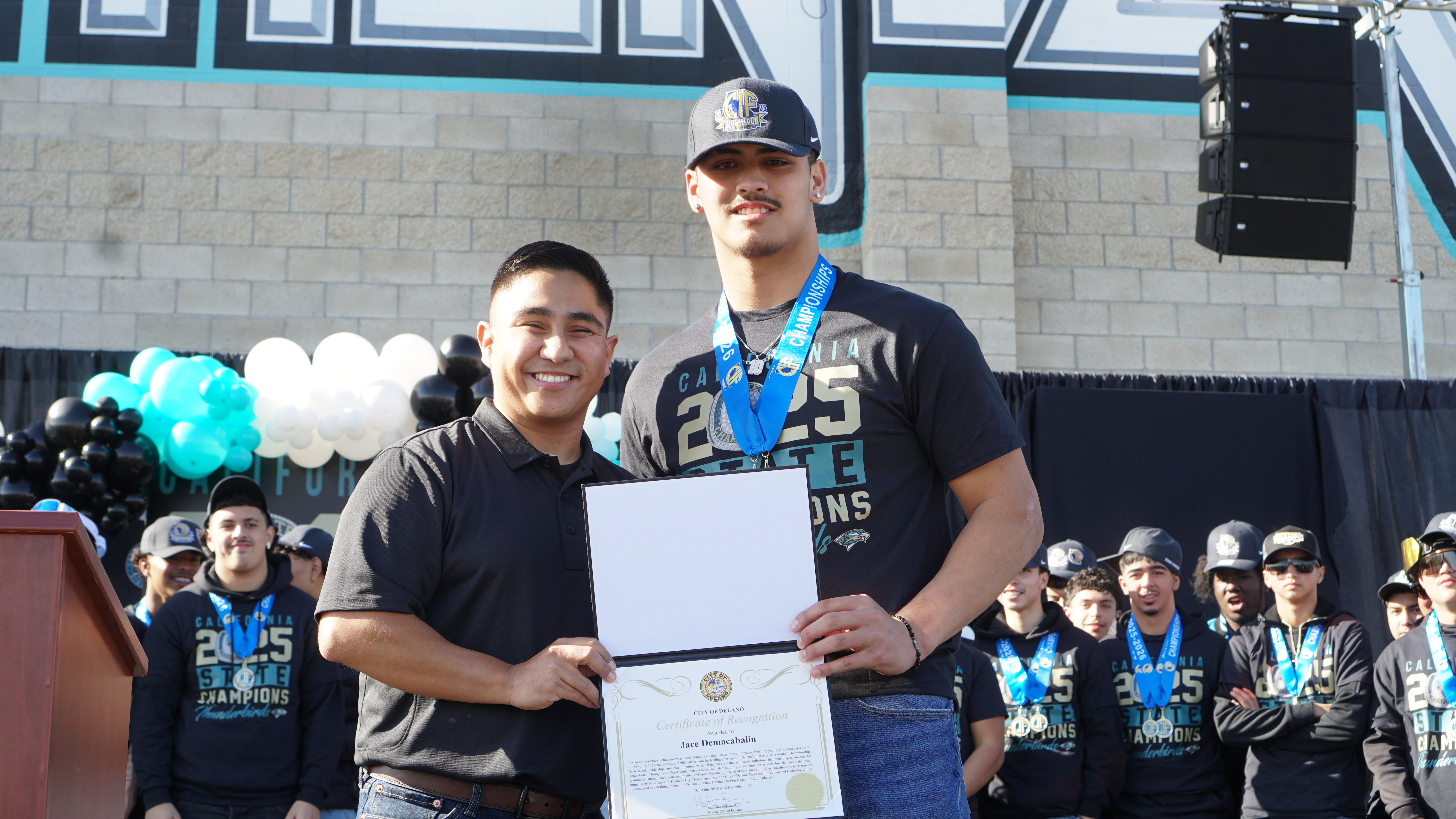 The image shows two people, one holding a certificate, standing in front of a crowd of people.