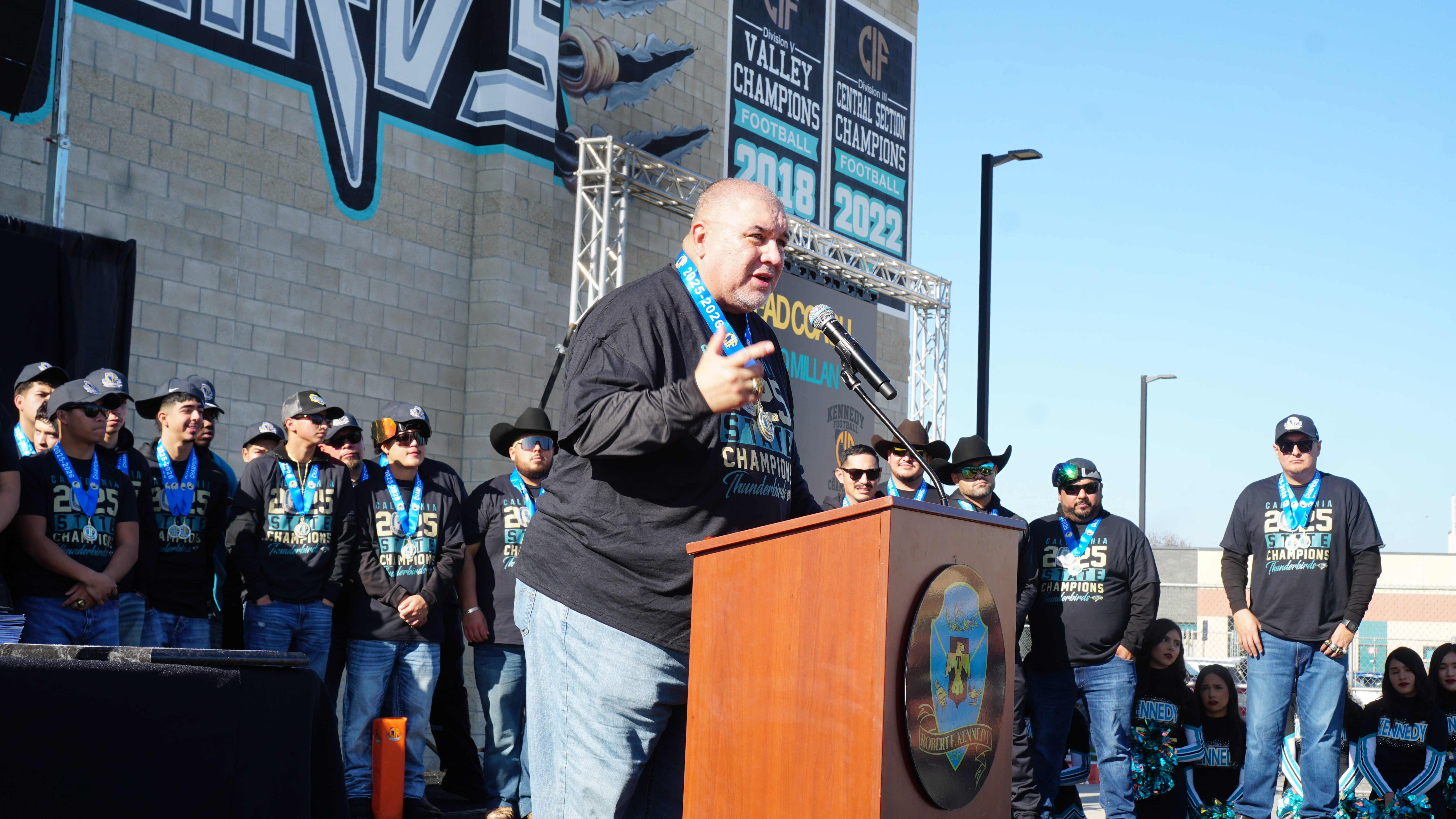 A man in a black suit is standing at a podium, addressing a crowd of people wearing matching shirts and jackets in the background.