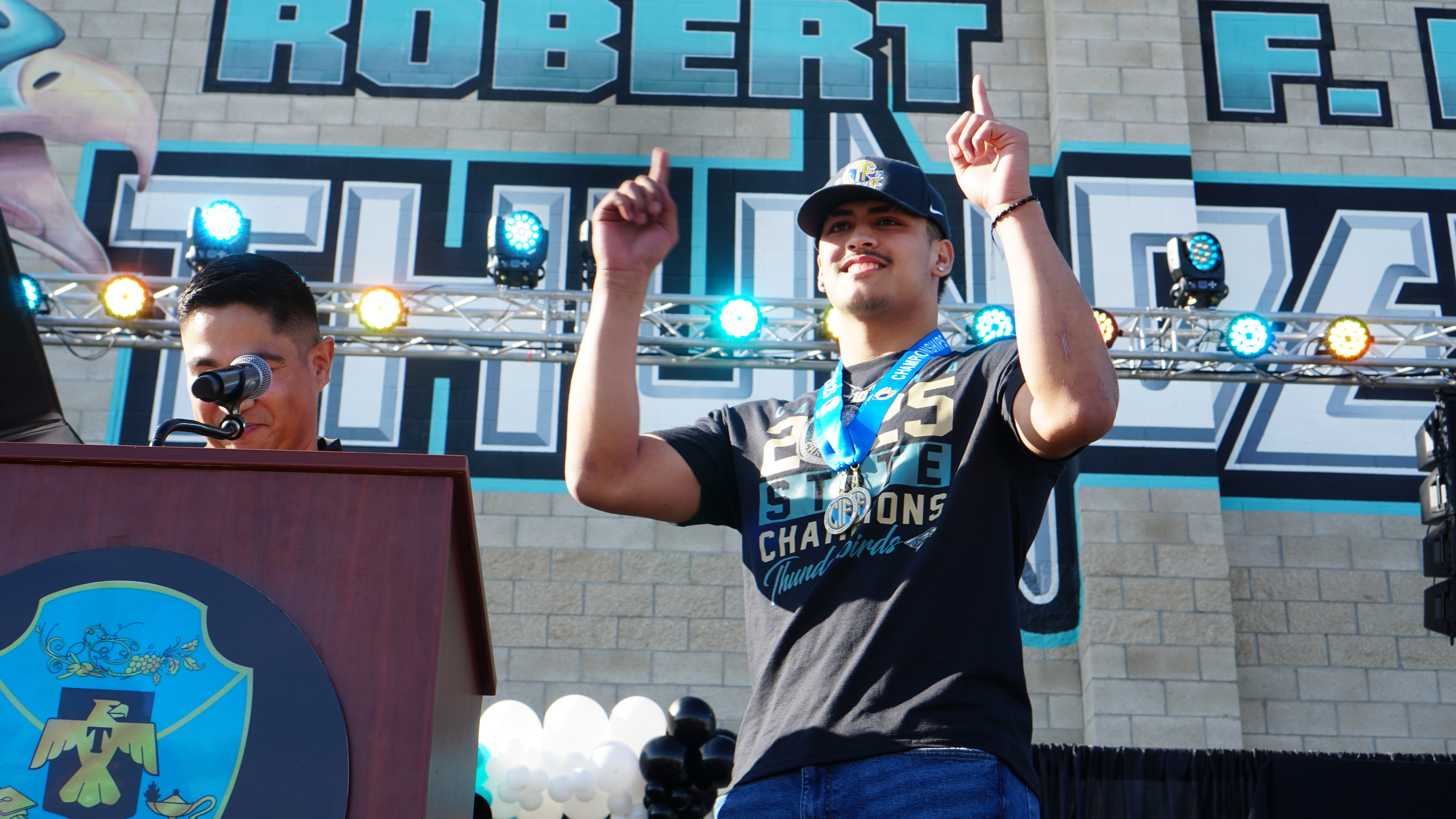 The image shows a person wearing a sports jersey and celebrating in front of a large sign that says "Robert F. Hall Champions".