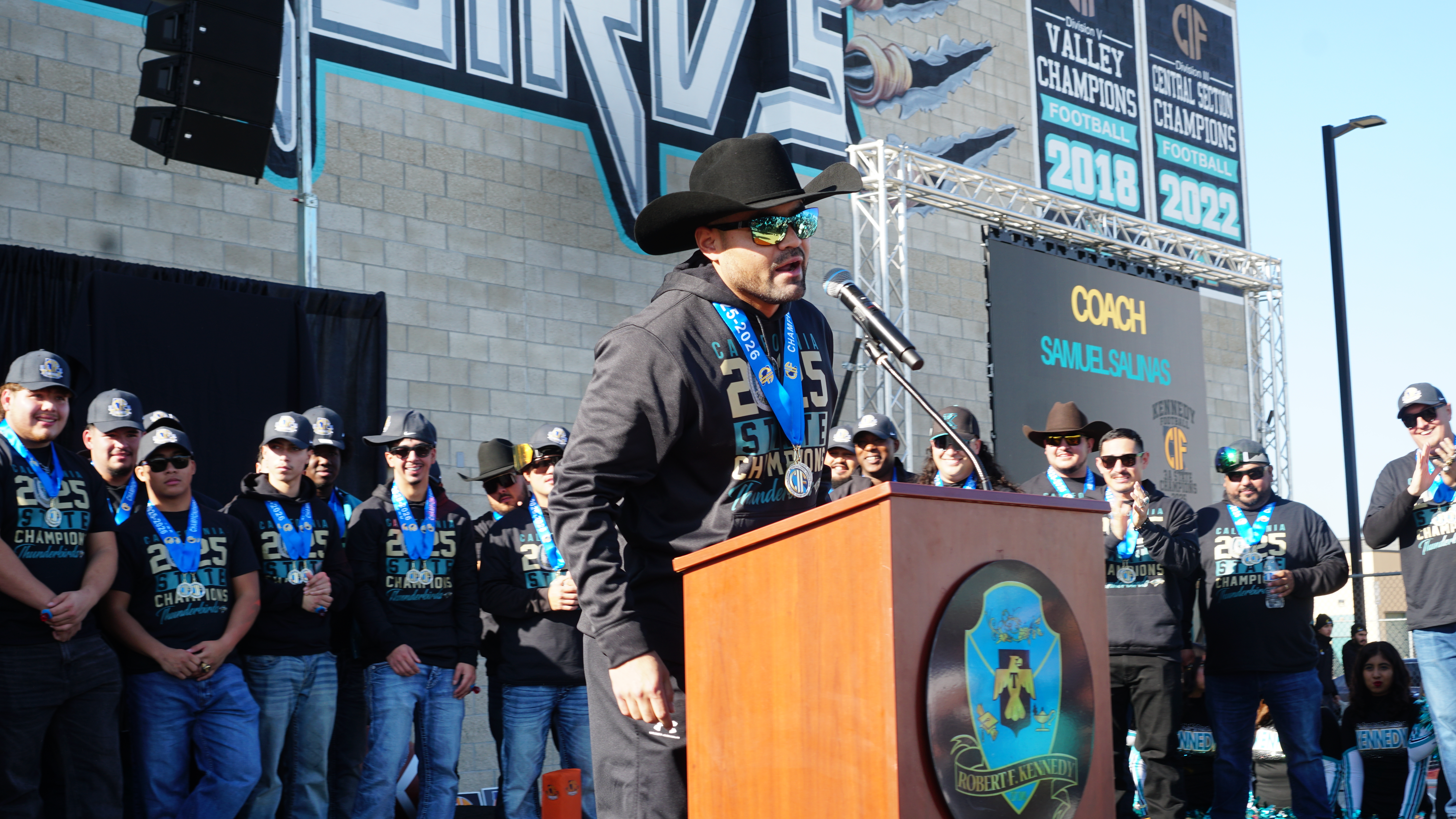 A man in a cowboy hat is standing at a podium, addressing a crowd of people wearing similar cowboy-style attire, with a large sign in the background.