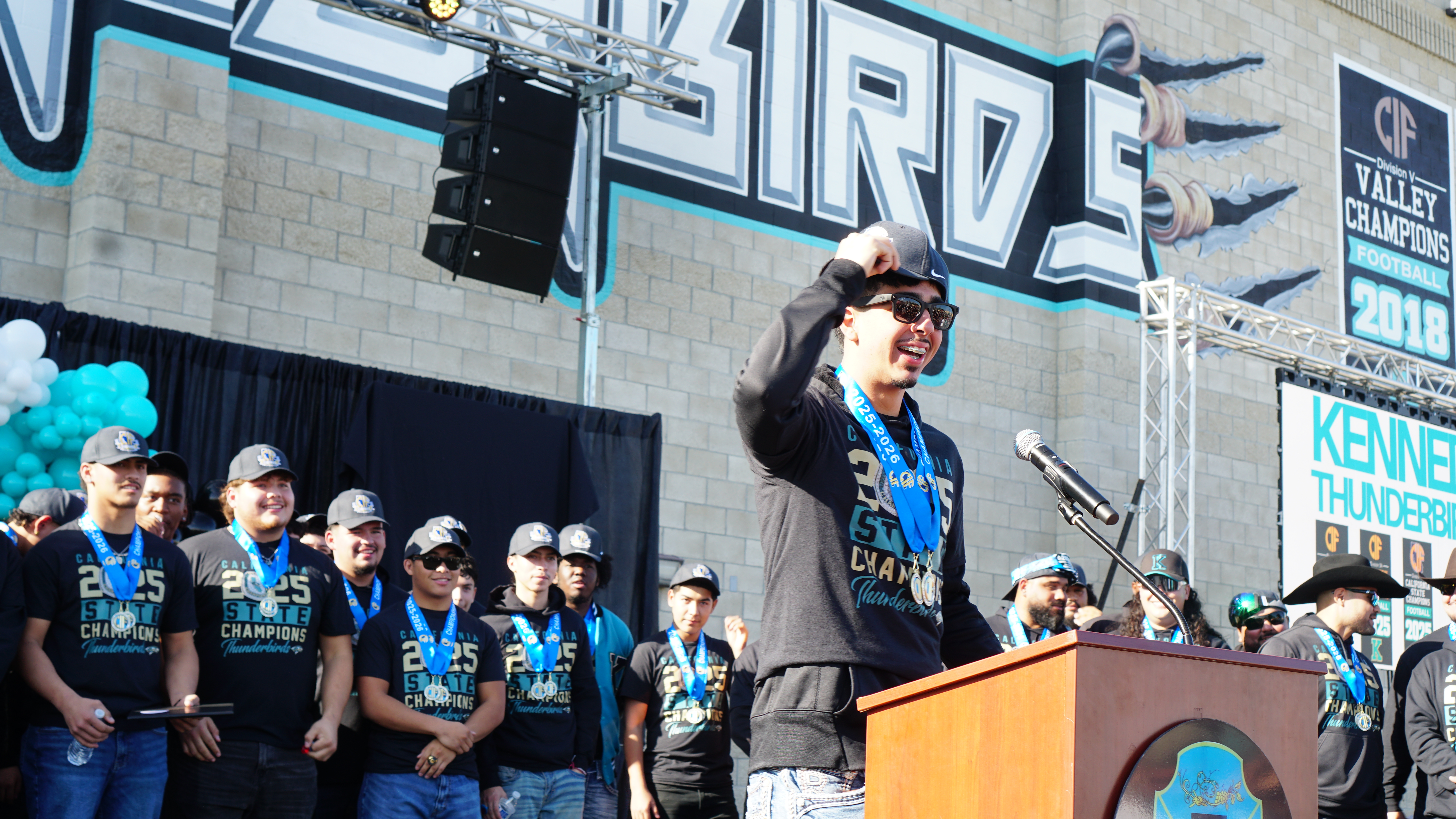 The image shows a large crowd of people gathered in front of a stage, with a person speaking into a microphone on the stage. The background features a large mural with the word "PRIDE" prominently displayed.