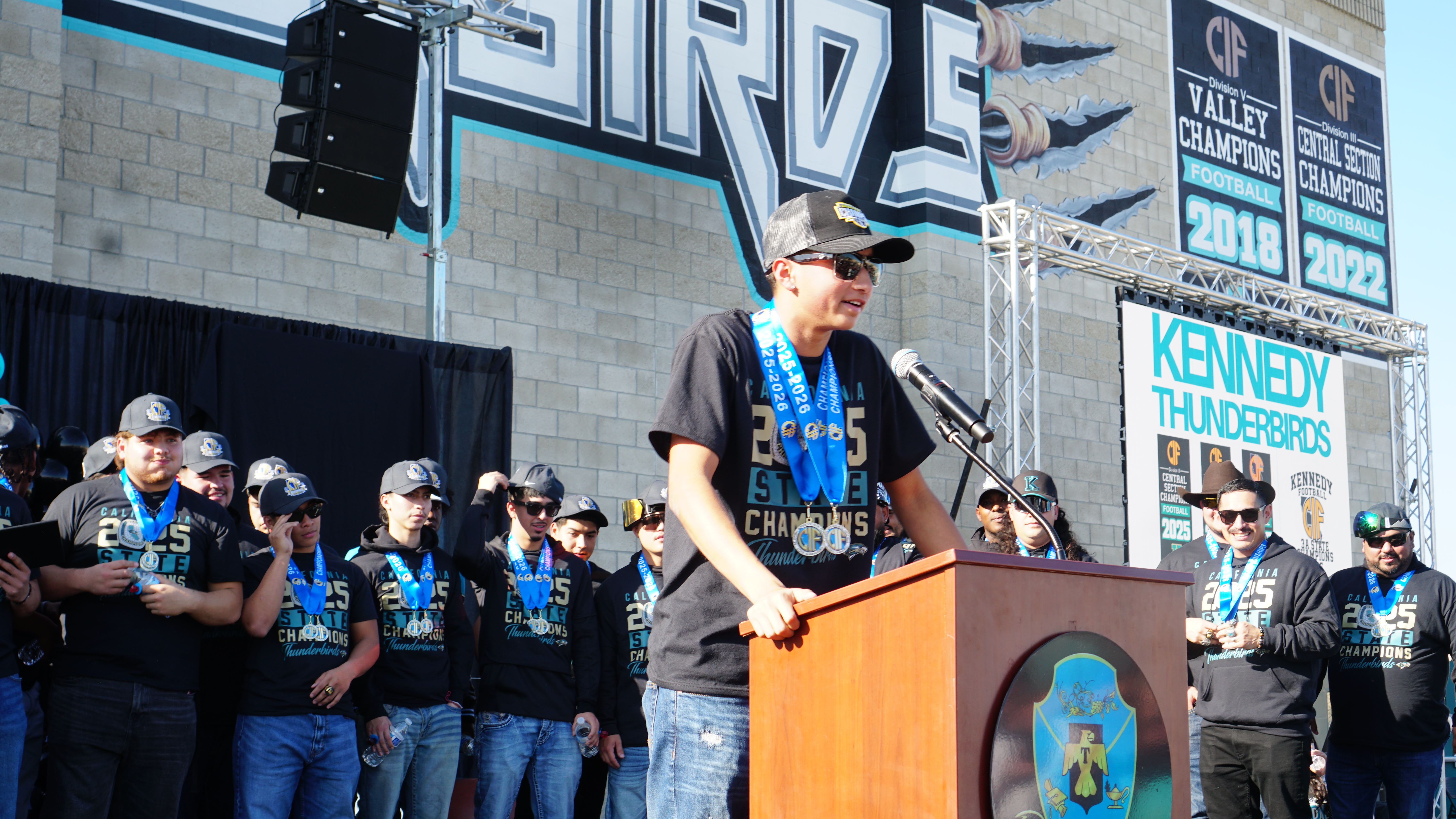 The image shows a large crowd of people gathered in front of a stage, with a person standing at a podium on the stage addressing the audience.