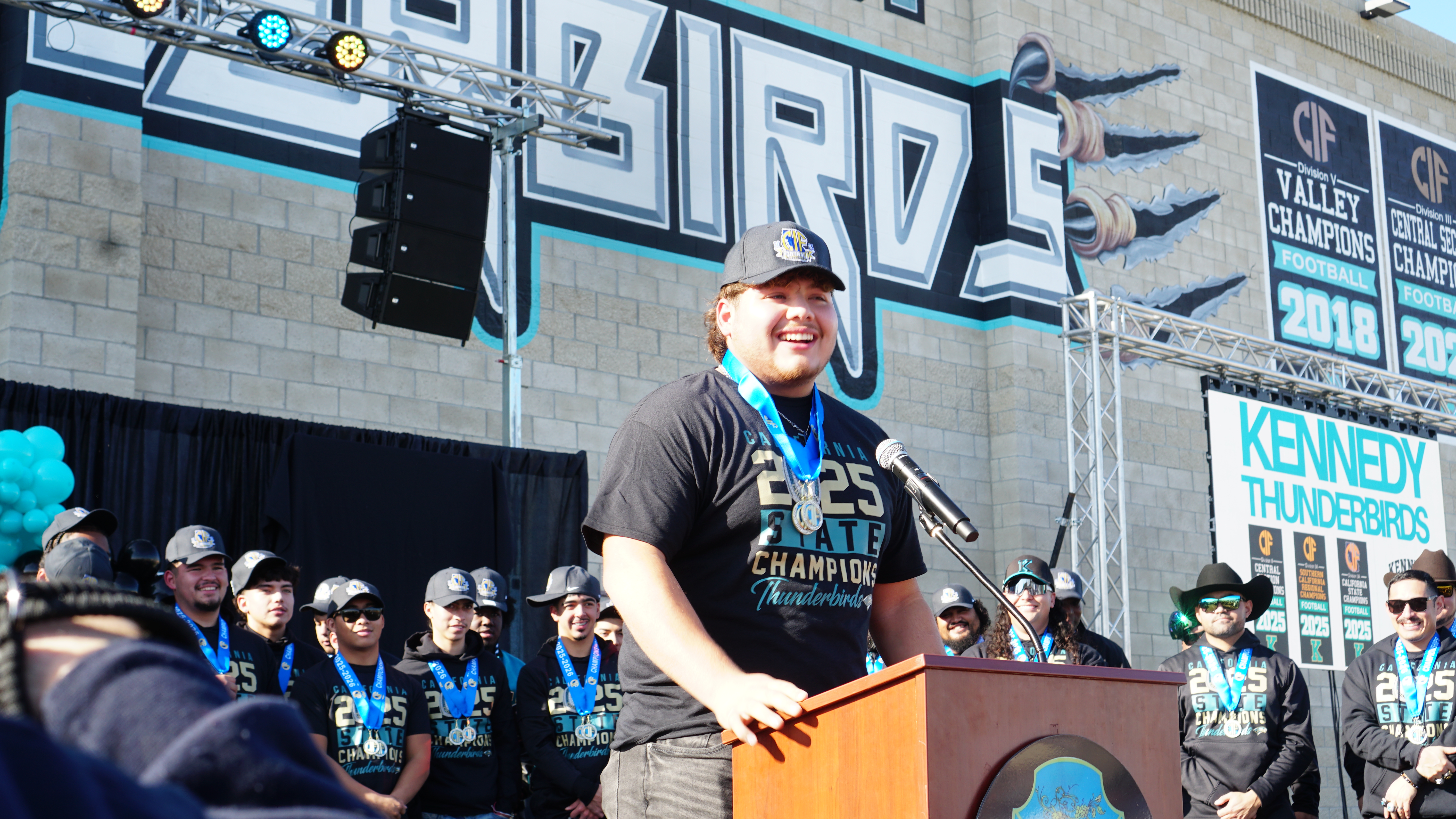 A person wearing a gray shirt and a medal stands at a podium in front of a crowd, with a large graphic backdrop displaying the event name.