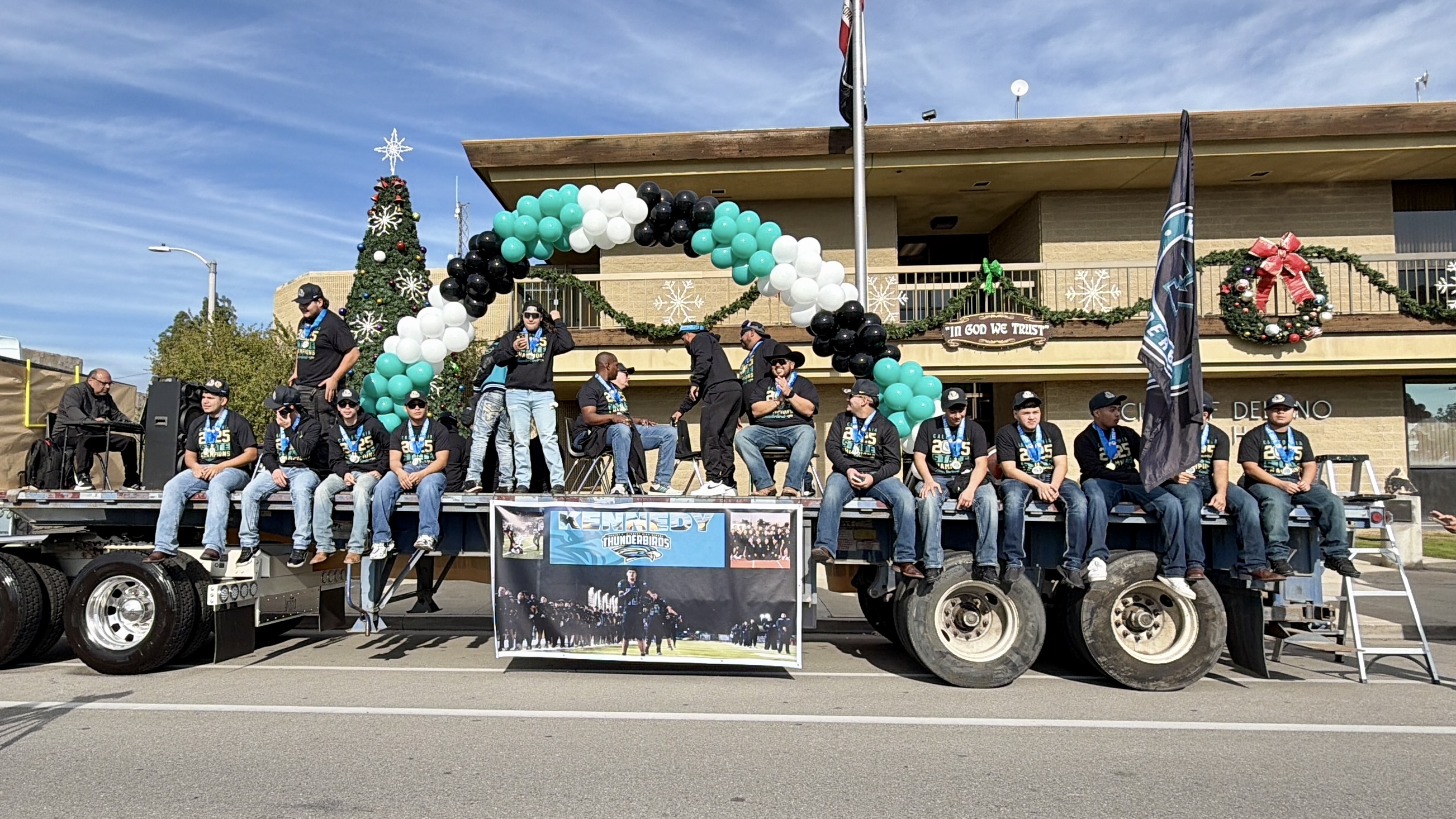 A group of people, likely members of an organization or club, are standing on the back of a large truck, surrounded by festive decorations and balloons in the foreground, with a building and a clear blue sky visible in the background.