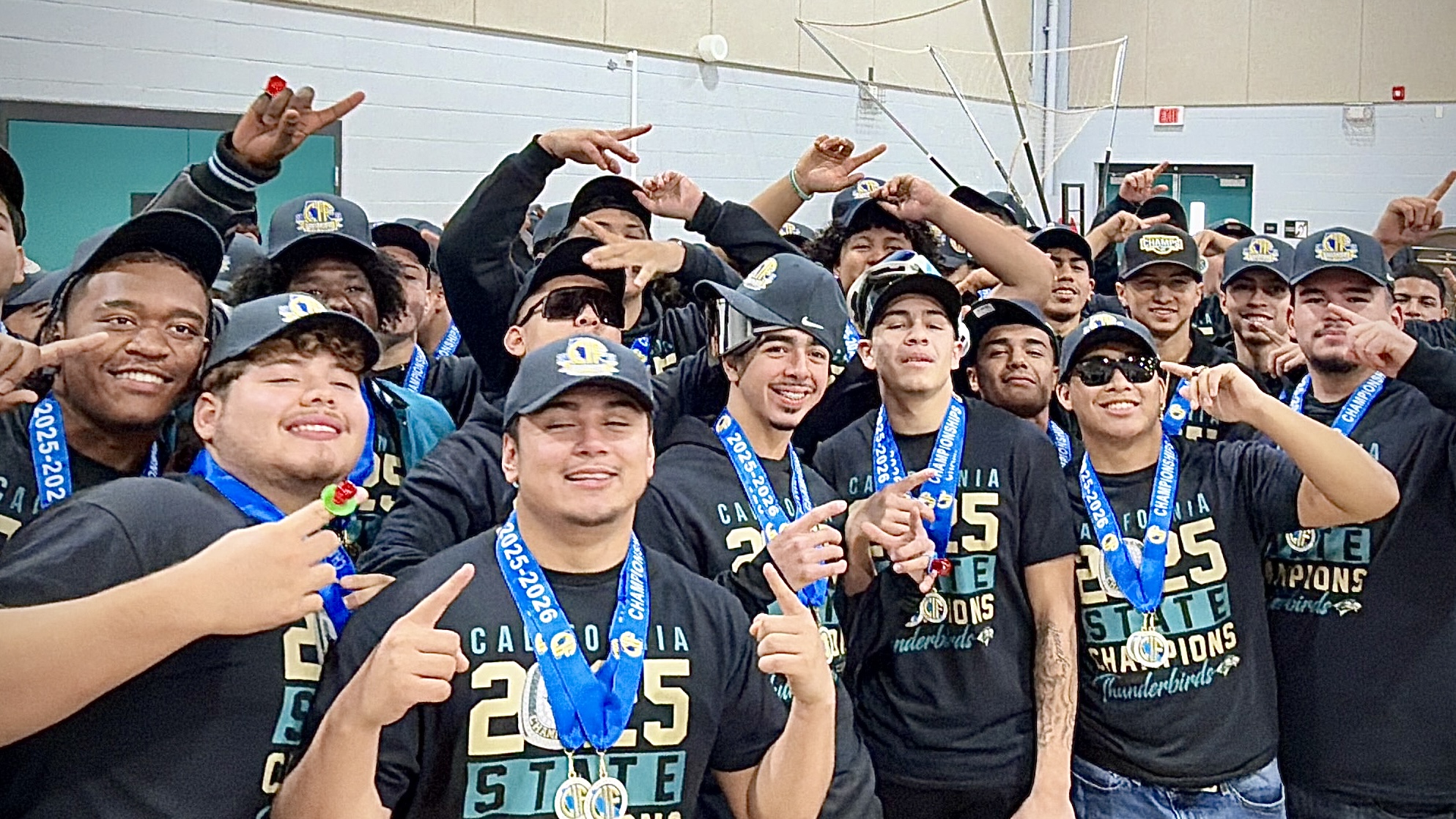 A group of enthusiastic people, mostly young adults, are gathered together and celebrating with their hands raised in the air, wearing matching team jerseys or shirts.