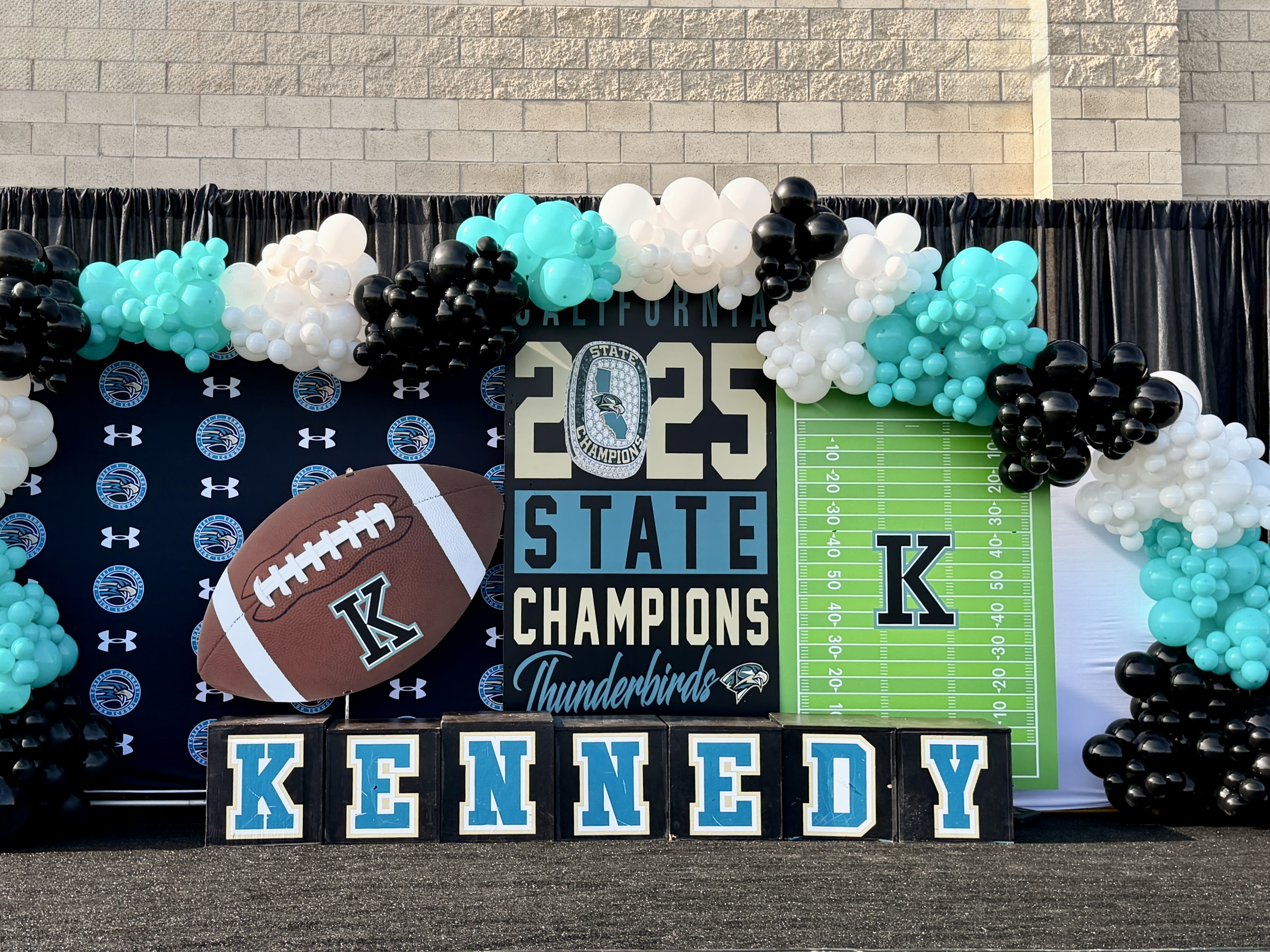 The image depicts a festive backdrop with a football-themed design, including a "State Champions" banner, a football, and the name "Kennedy" displayed prominently. The background is adorned with colorful balloons in shades of turquoise, white, and black, creating a celebratory atmosphere.