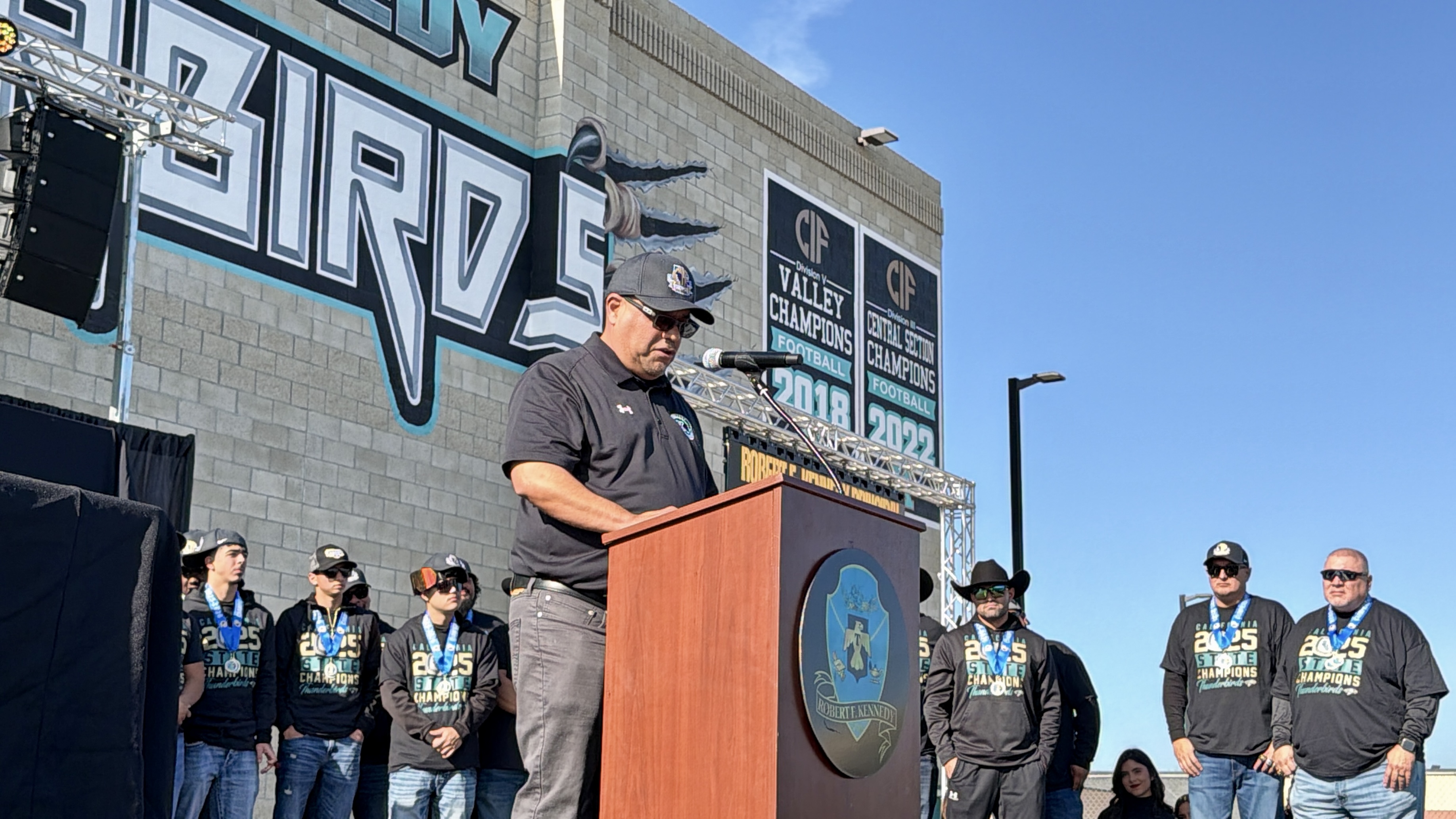 A man is standing at a podium in front of a large mural, surrounded by a group of people wearing matching uniforms.