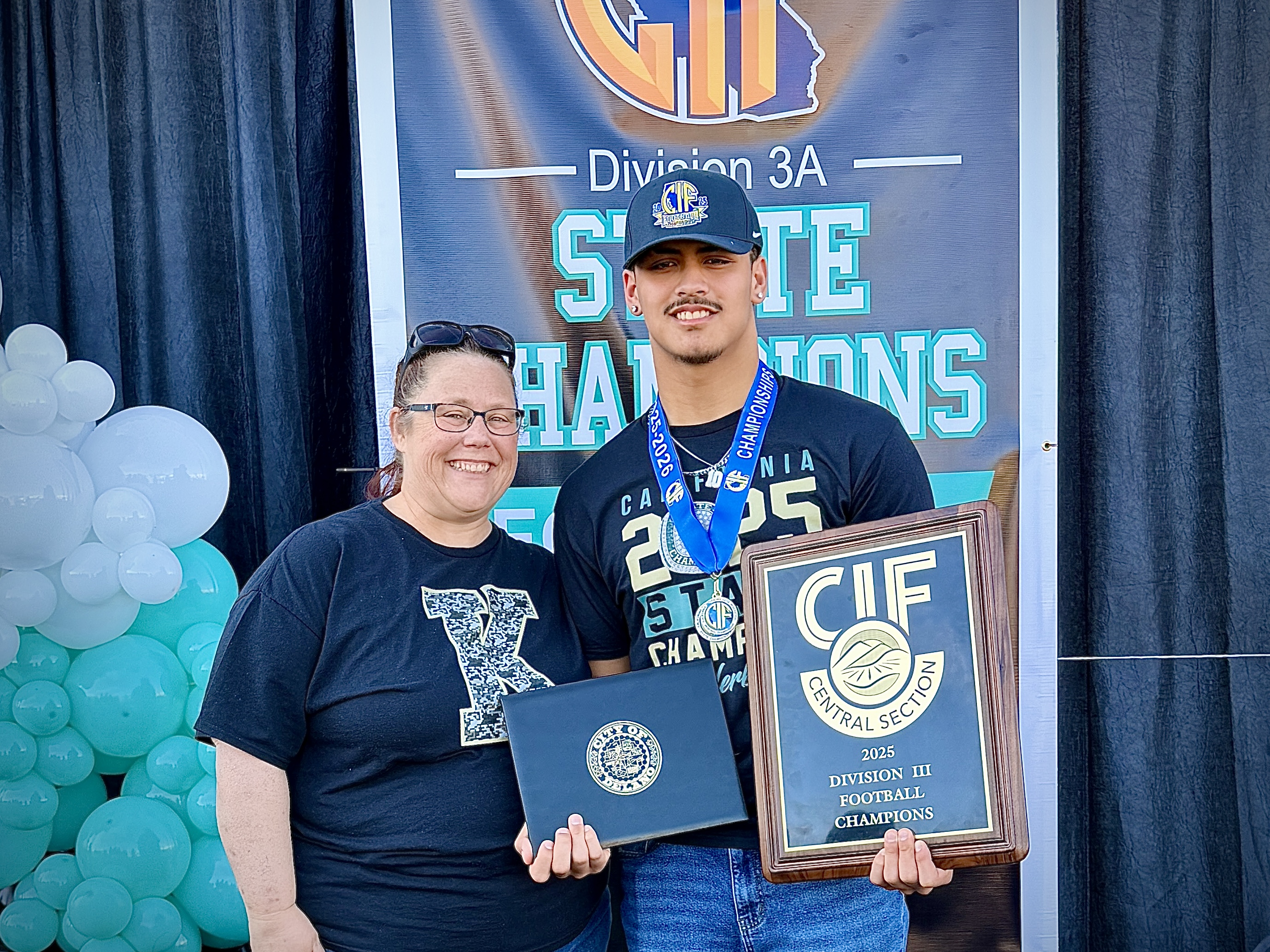 Two individuals, a man and a woman, are standing in front of a banner that displays the text "Division 3A State Champions". The man is holding a trophy, and the woman is holding a framed certificate. The background appears to be a celebration or event setting, with balloons and other decorations visible.