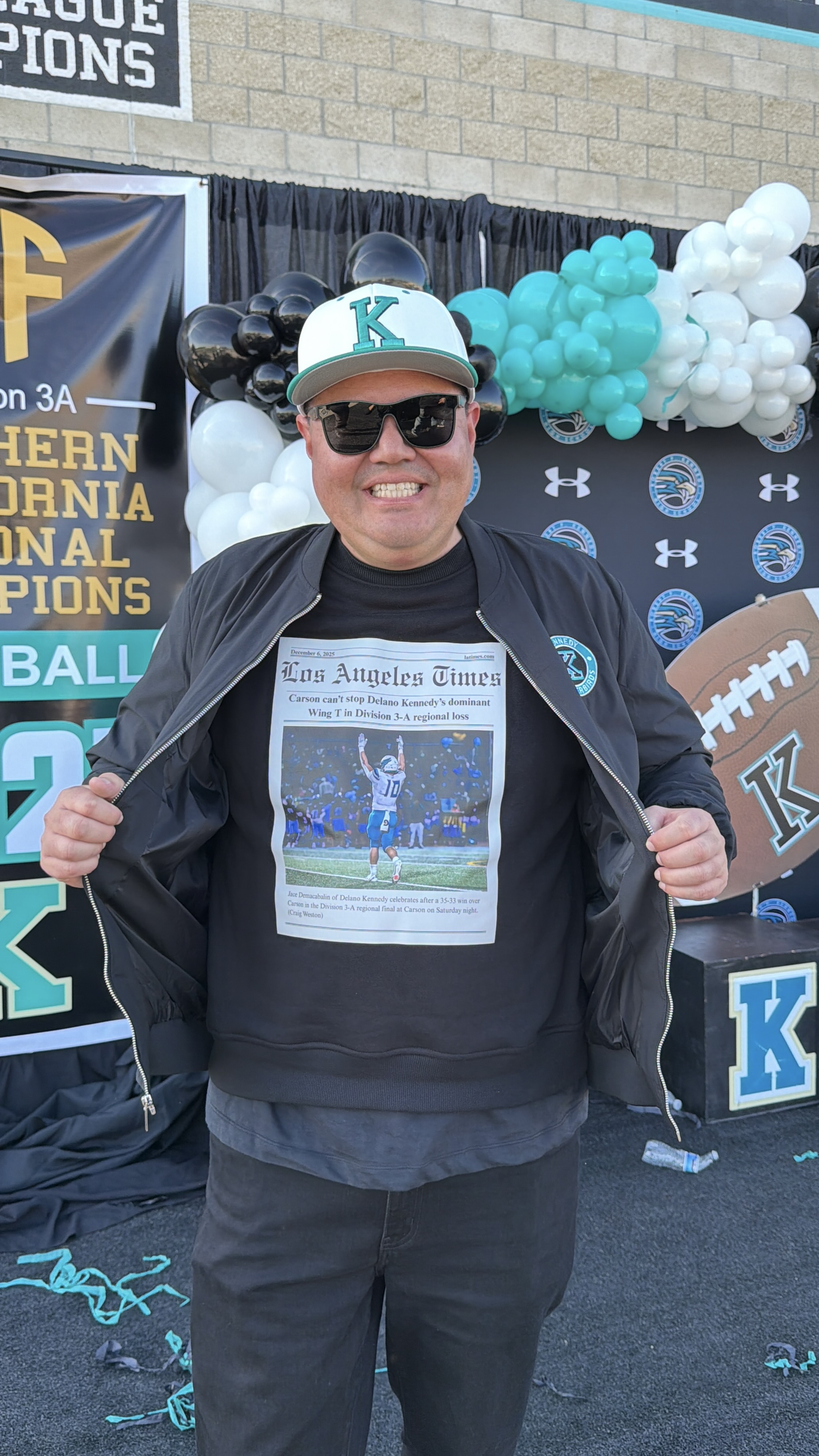 A smiling person wearing a black jacket and a cap with the letter "K" stands in front of a backdrop featuring sports-related banners and decorations.