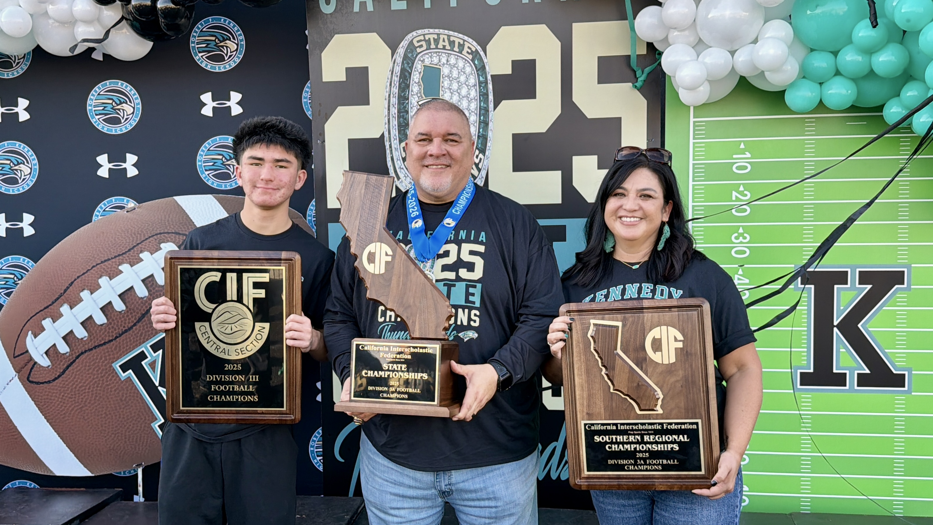 The image shows three people holding various awards and trophies, with a football-themed background featuring balloons and a football field.