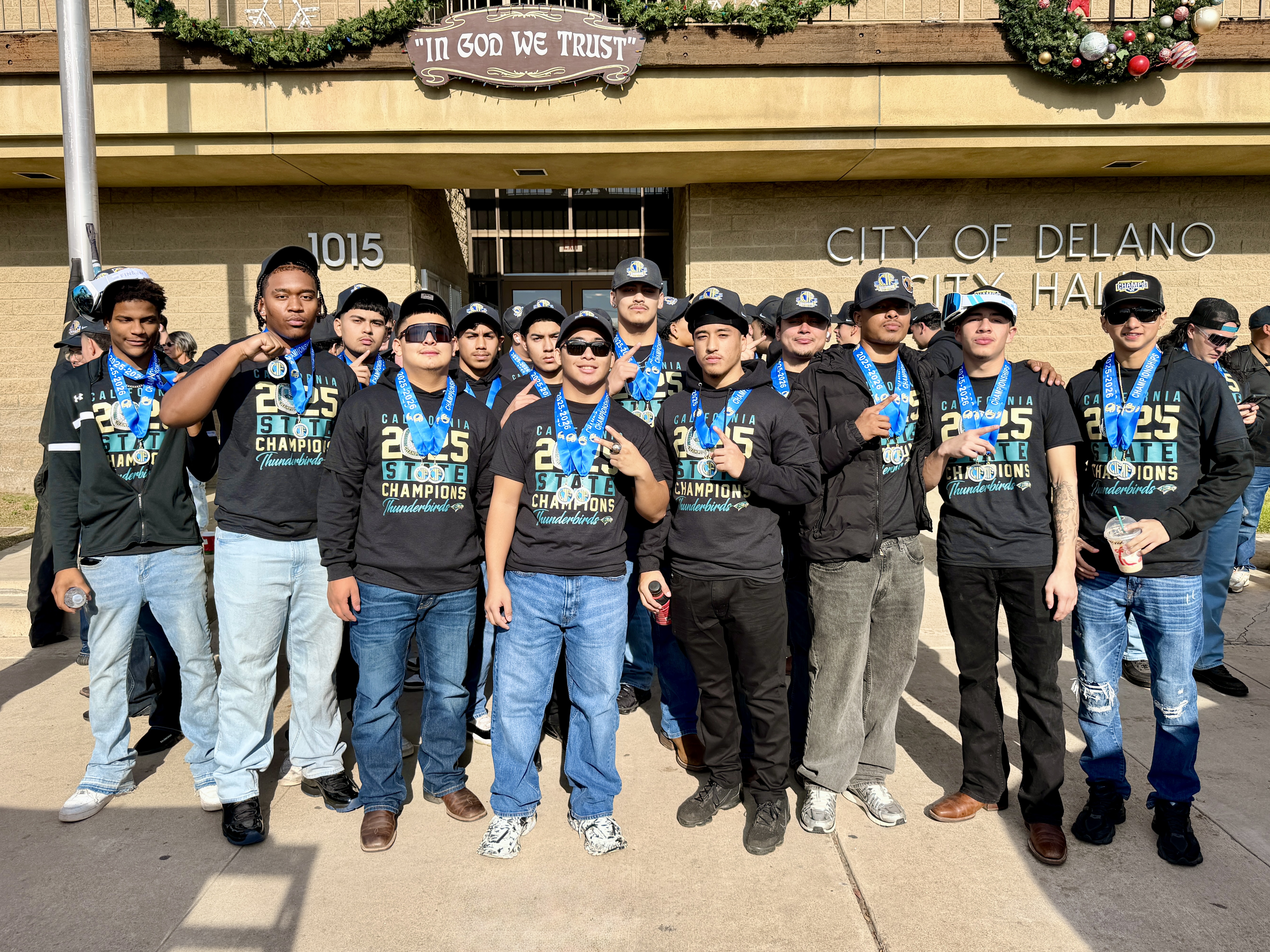 A group of people, likely a sports team or club, posing together in front of a building with a sign that says "City of Delano".