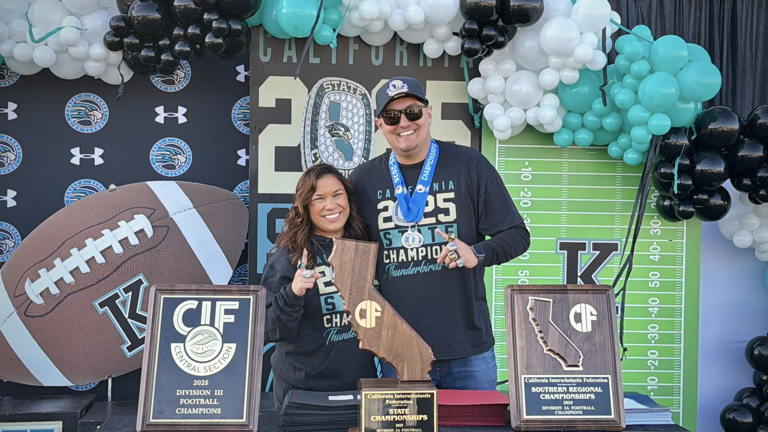 The image shows a couple posing in front of a festive backdrop featuring sports-themed decorations, including a large football and a football field design.