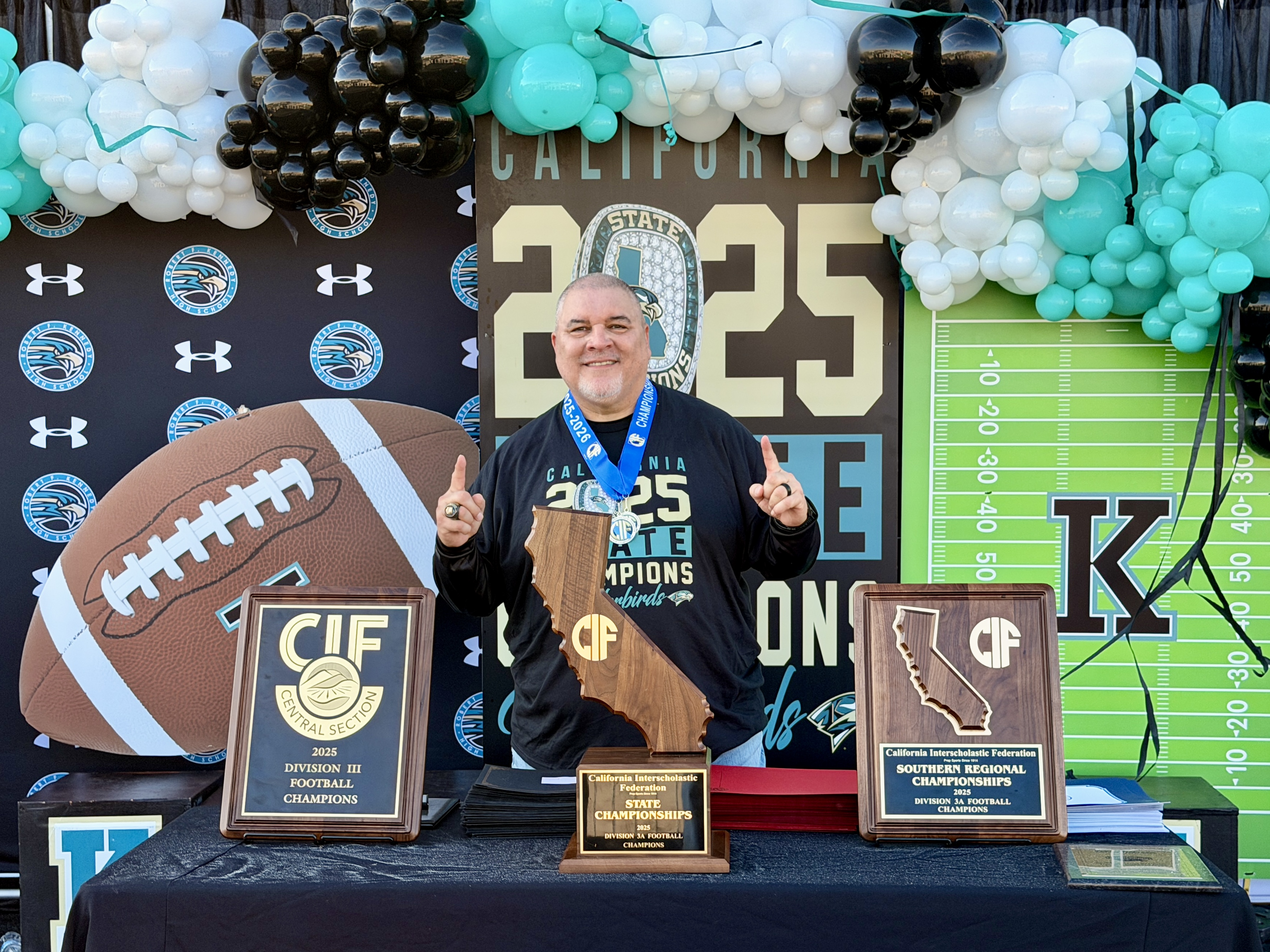 A man stands in front of a sports-themed backdrop, holding several trophies and awards.