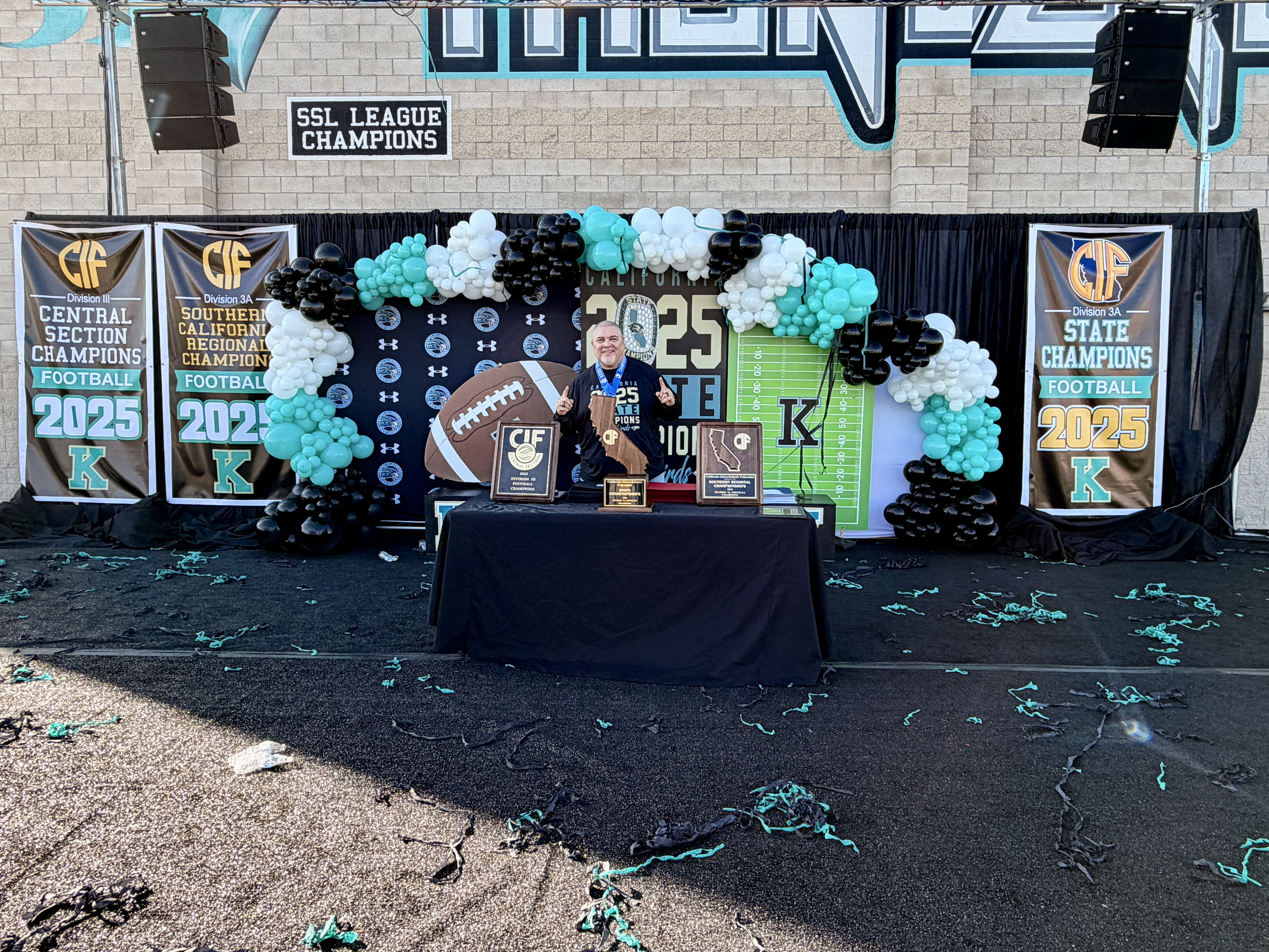The image depicts a festive celebration setup with a stage, banners, and balloon decorations, likely for a championship or graduation event.