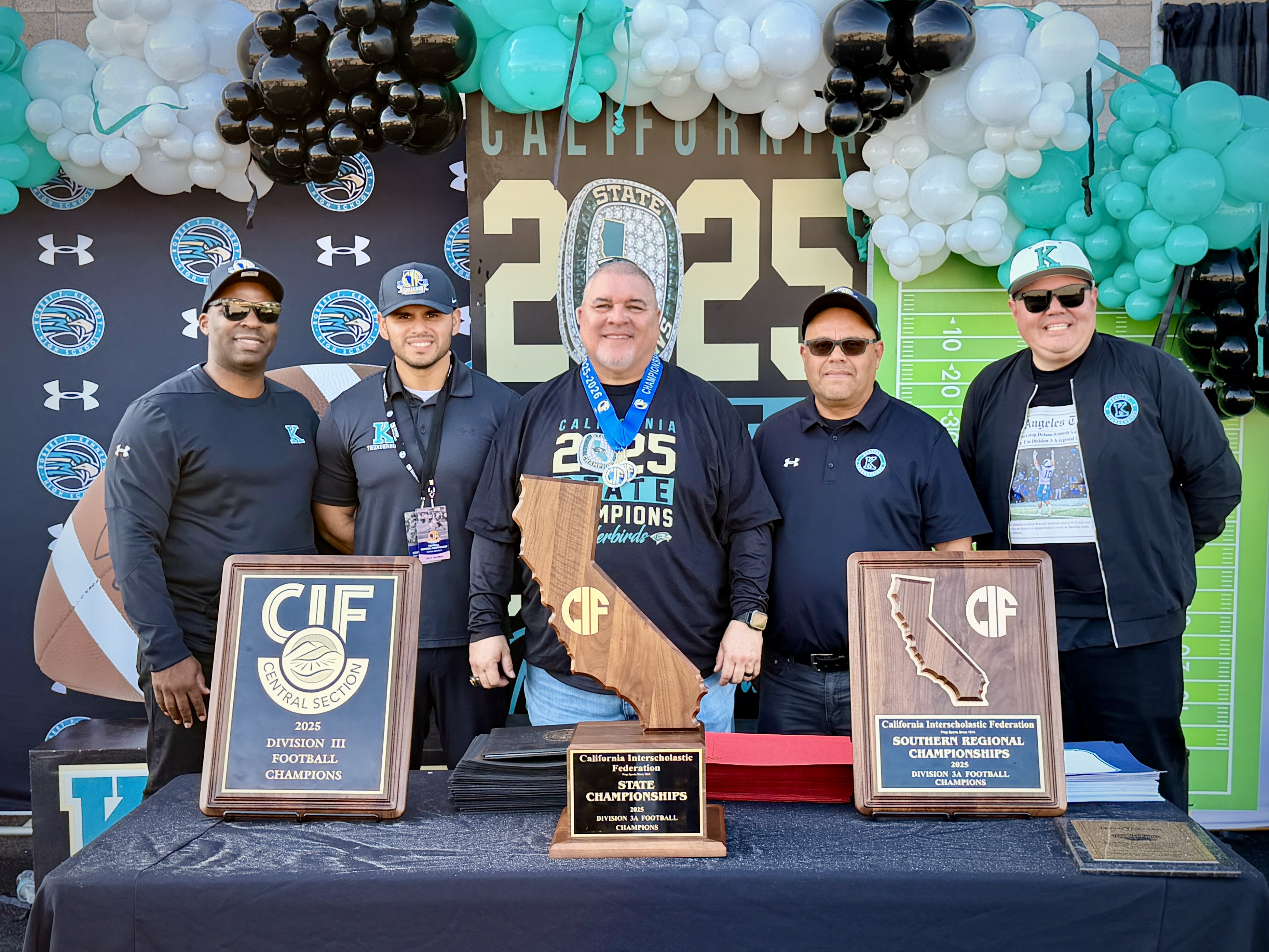 The image shows a group of people standing behind a table displaying various awards and trophies, with a backdrop of colorful balloons and decorations.