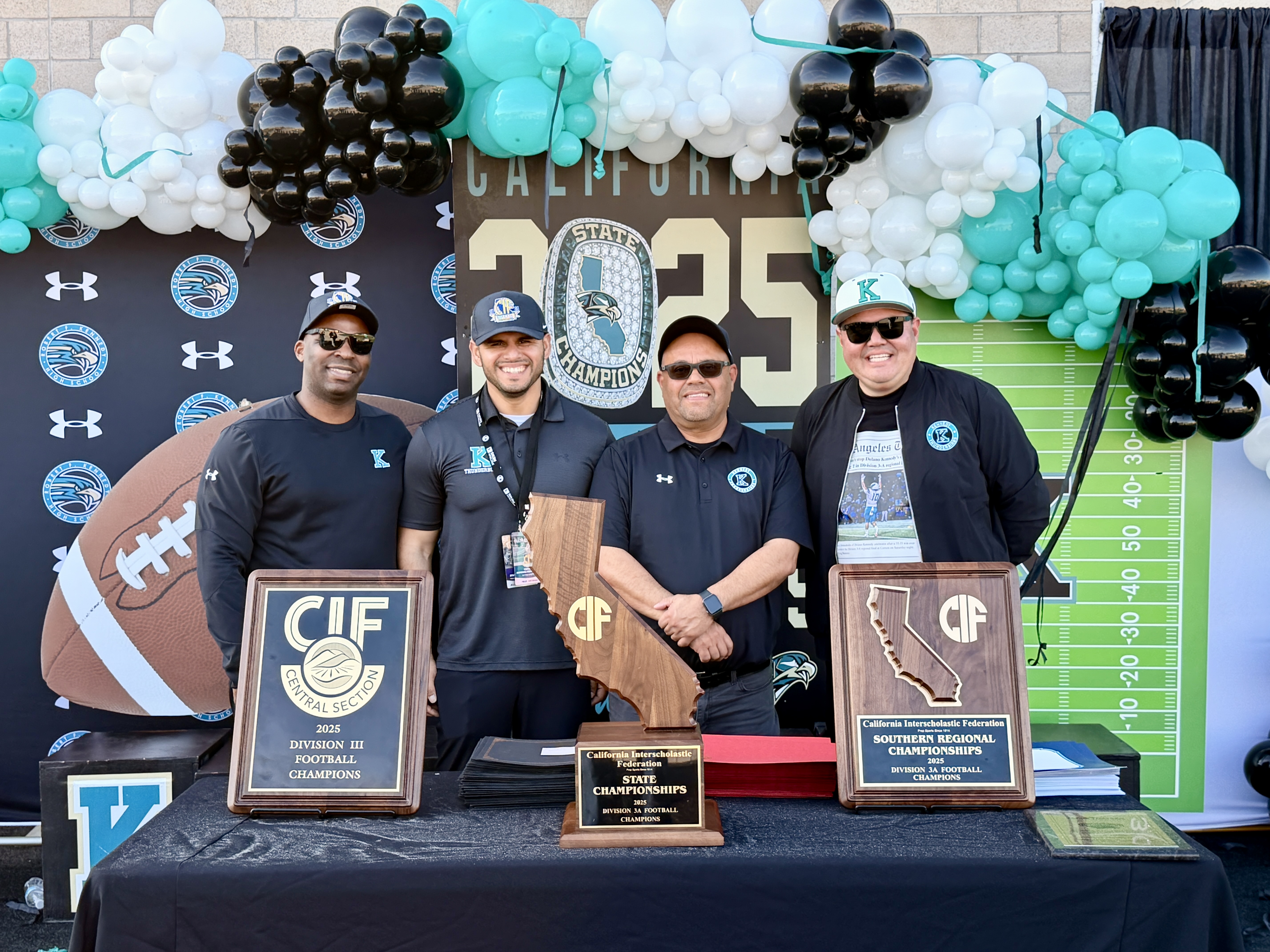 The image shows a group of four individuals posing in front of a backdrop featuring a football-themed design, including balloons, a scoreboard, and a football field. They appear to be holding awards or trophies, suggesting they are being recognized for some achievement.