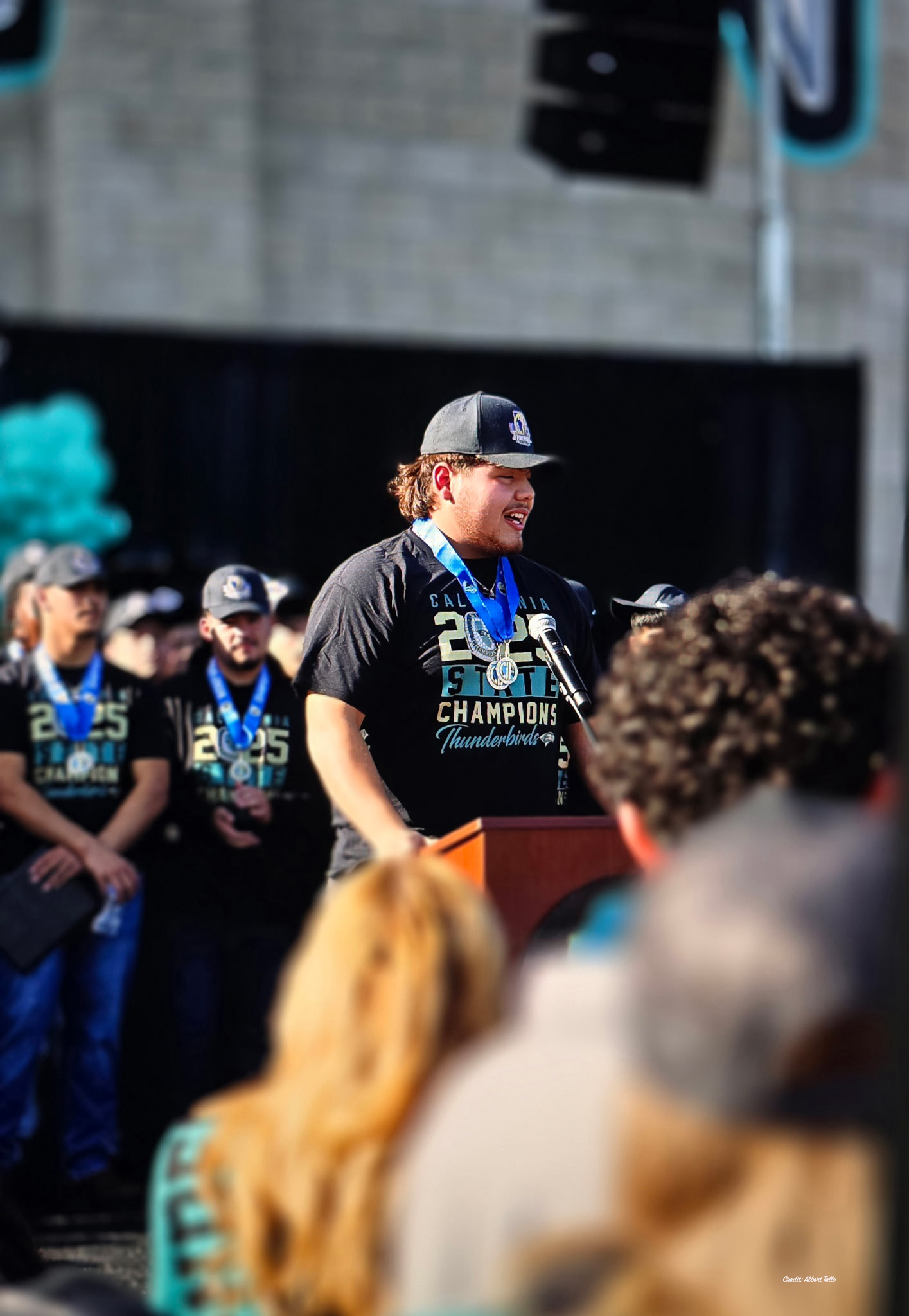 A man wearing a "Champion" hat and shirt is standing in front of a crowd, addressing them from a podium.