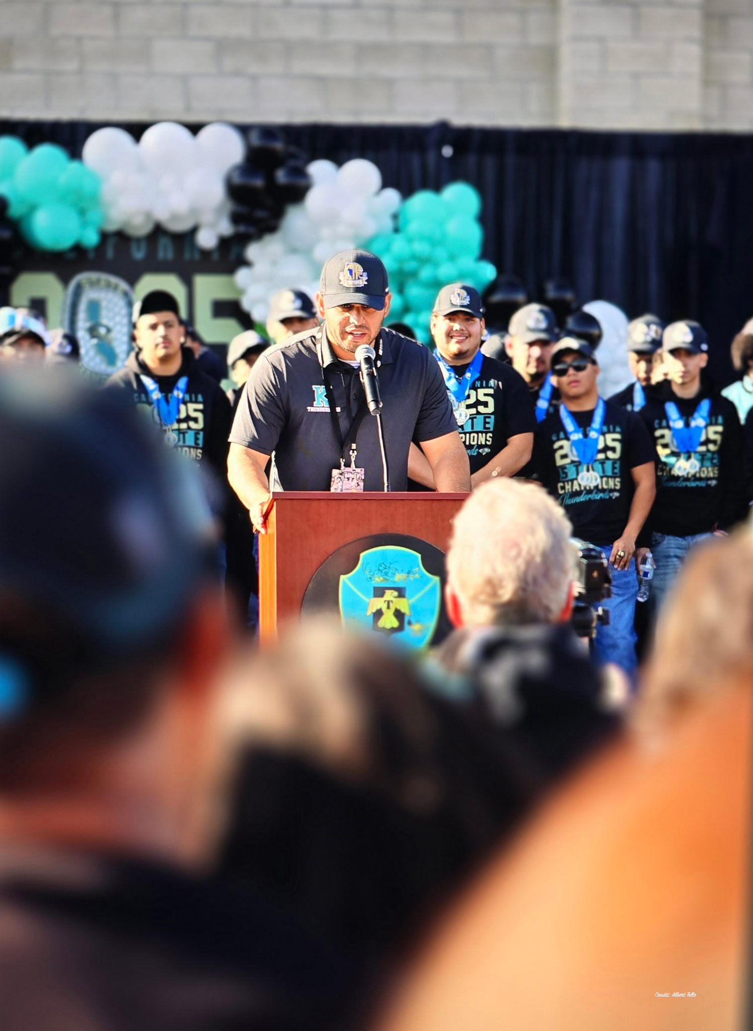 A man in a black shirt and cap is standing at a podium, addressing a crowd of people wearing matching blue shirts and hats, with colorful balloons decorating the background.