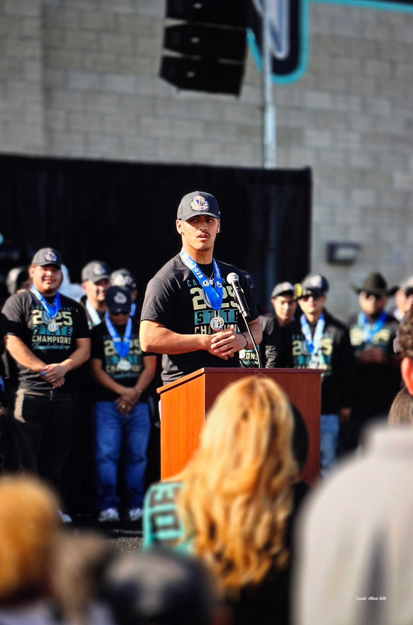 A man in a black shirt and cap is standing at a podium, addressing a crowd of people in the background.