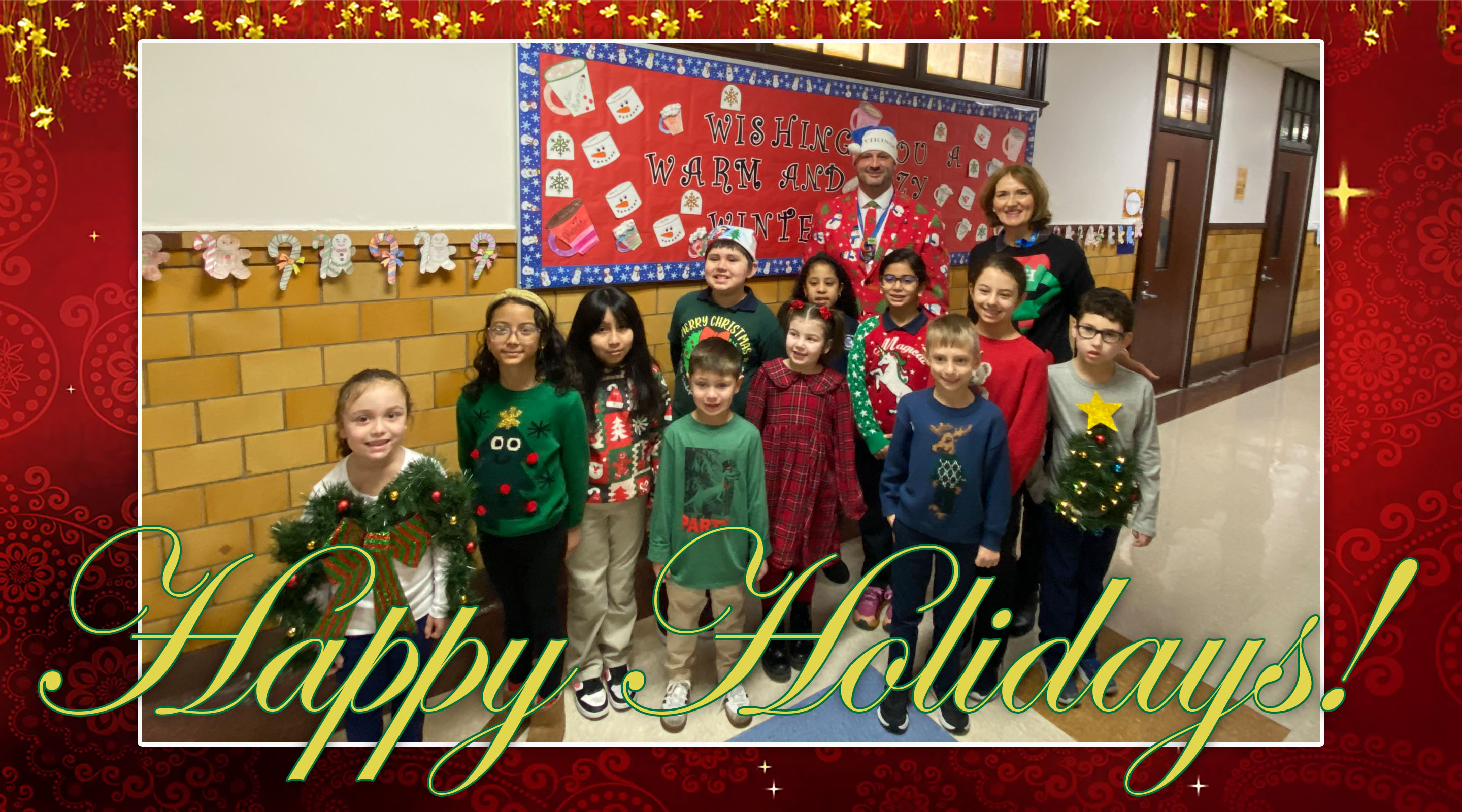 A group of children and two adults pose for a holiday photo in front of a festive classroom bulletin board.