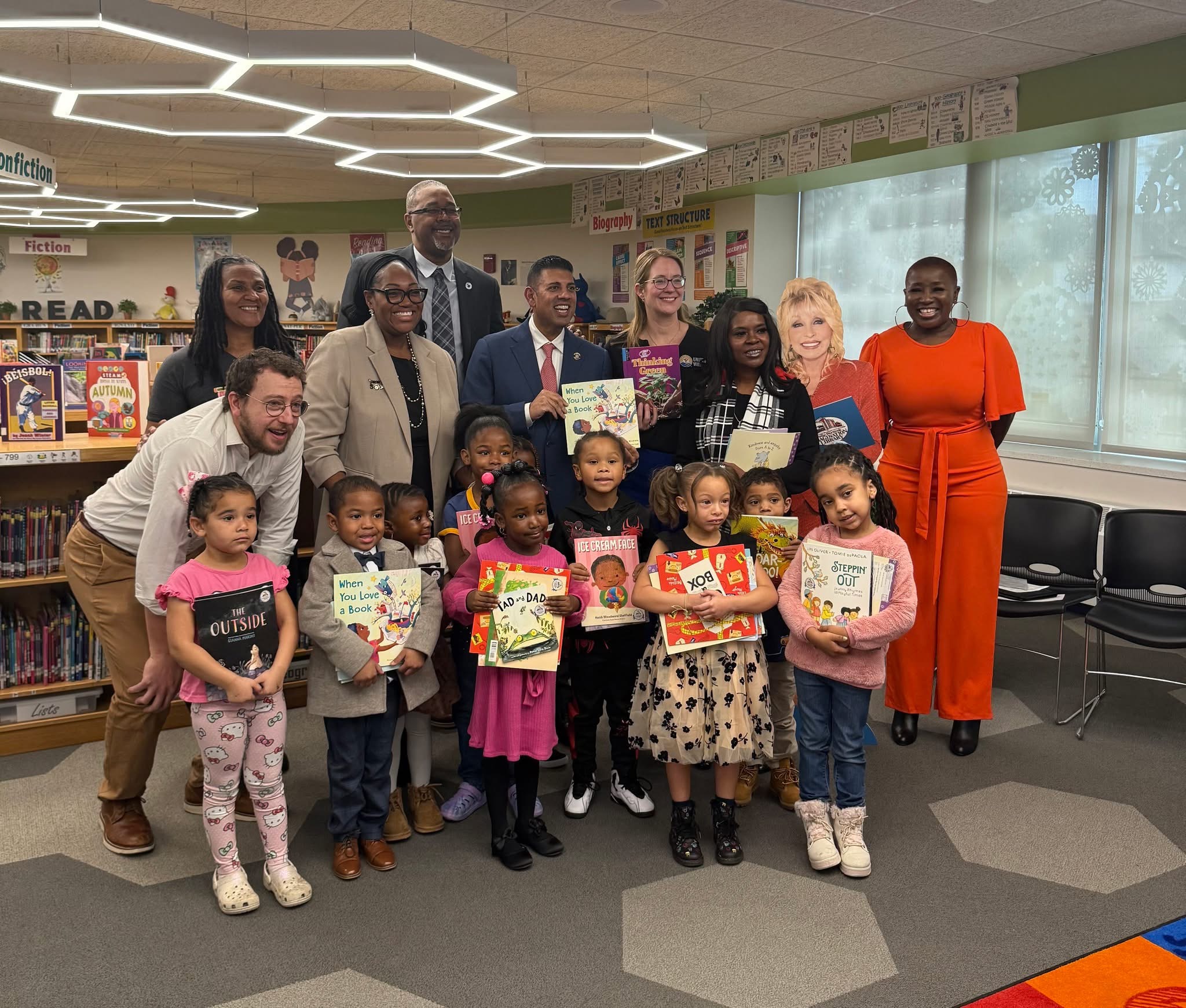 A group of adults and children, some in formal attire and others in casual clothing, are gathered in what appears to be a school or community center setting, surrounded by bookshelves and educational materials.