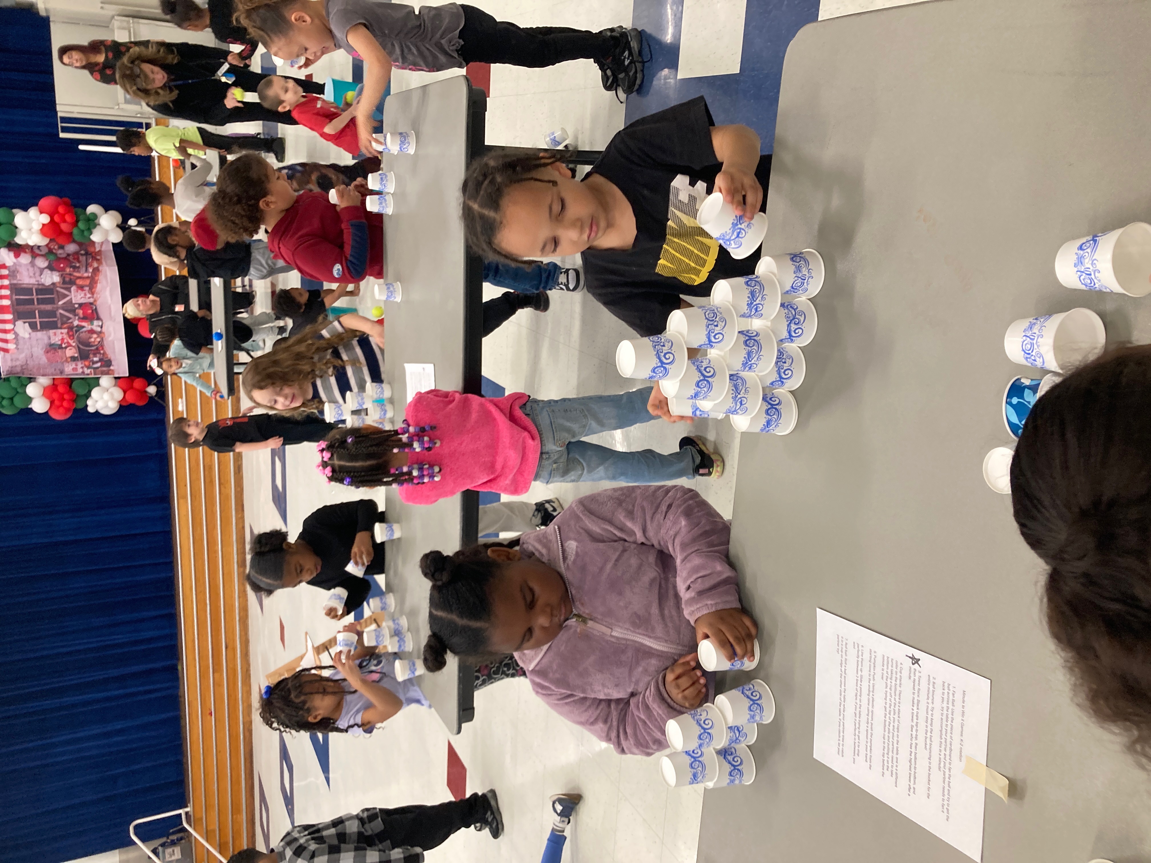 A group of children sitting at tables, engaged in an activity involving cups and other materials, with a colorful backdrop and adults present in the background.