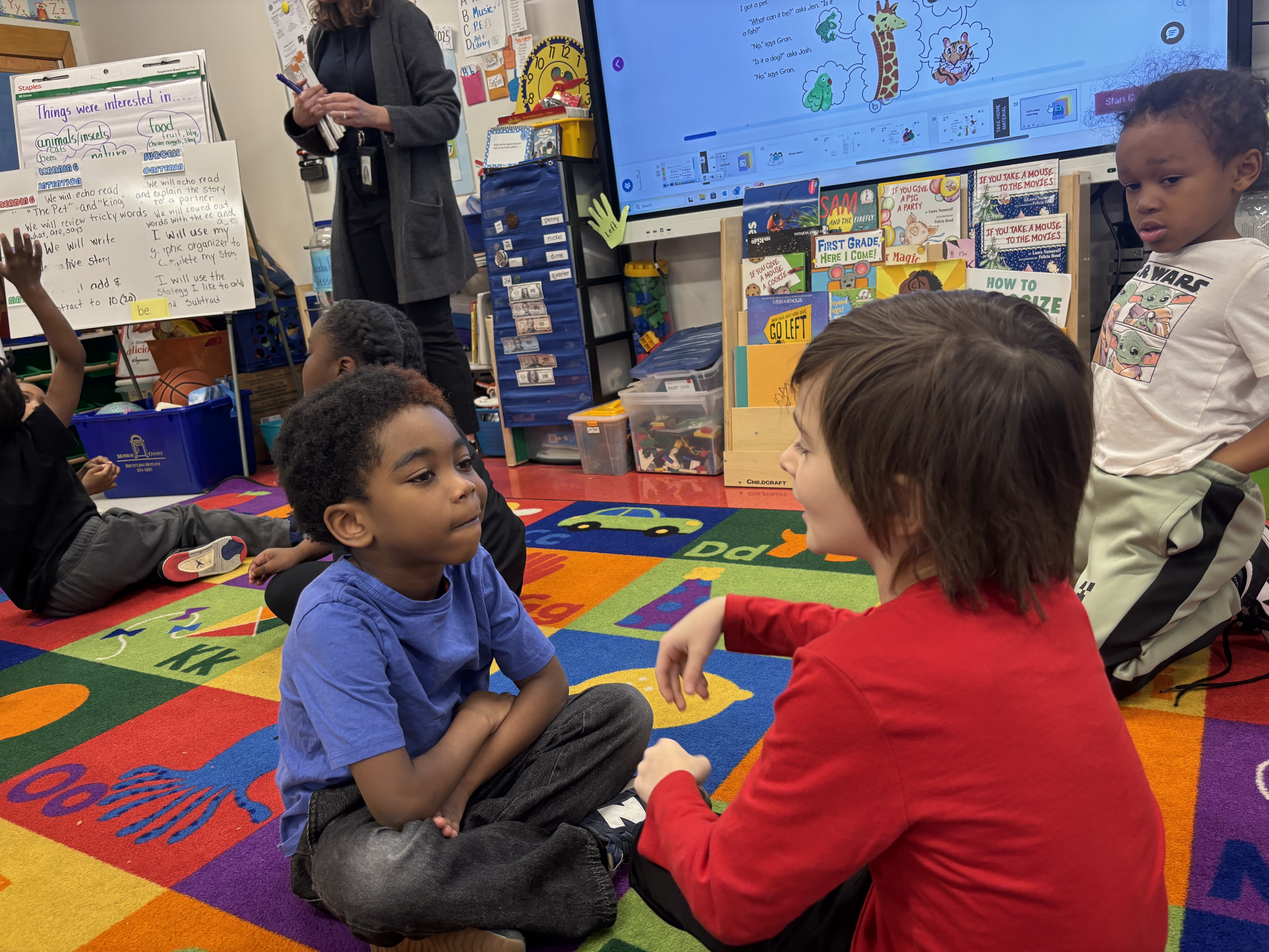 The image shows a classroom setting with a group of young children sitting on a colorful rug, engaged in what appears to be a discussion or activity led by an adult figure standing in the background.