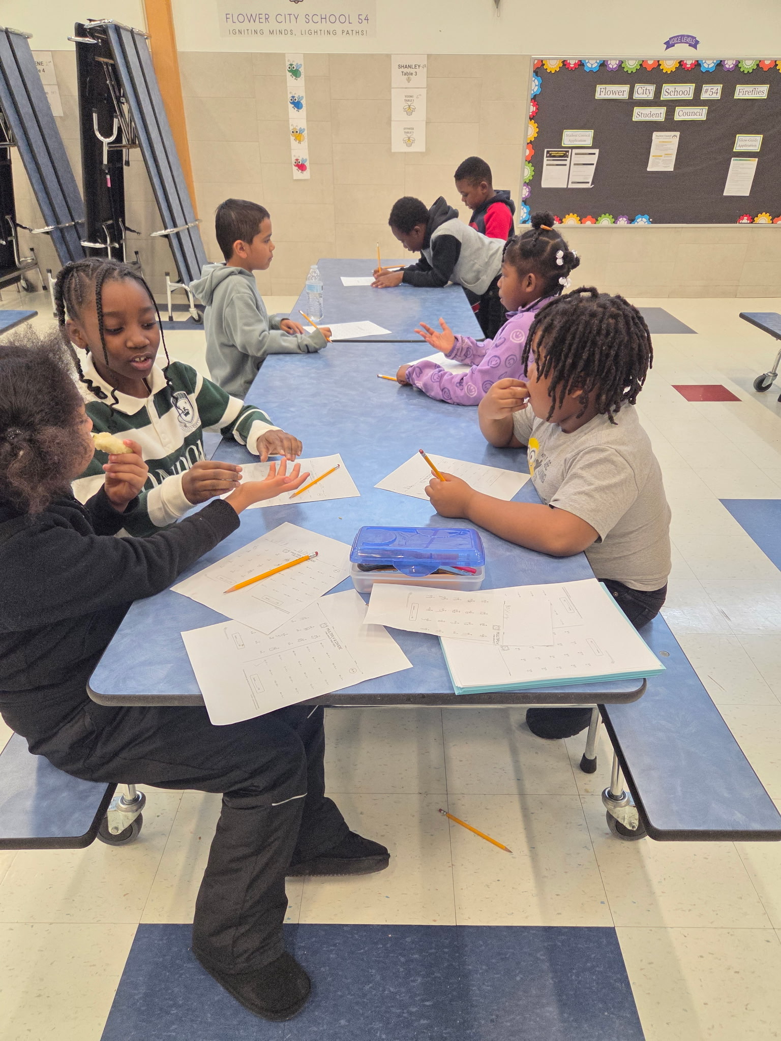 A group of young students sitting at desks in a classroom, engaged in various learning activities, with educational materials and displays visible in the background.