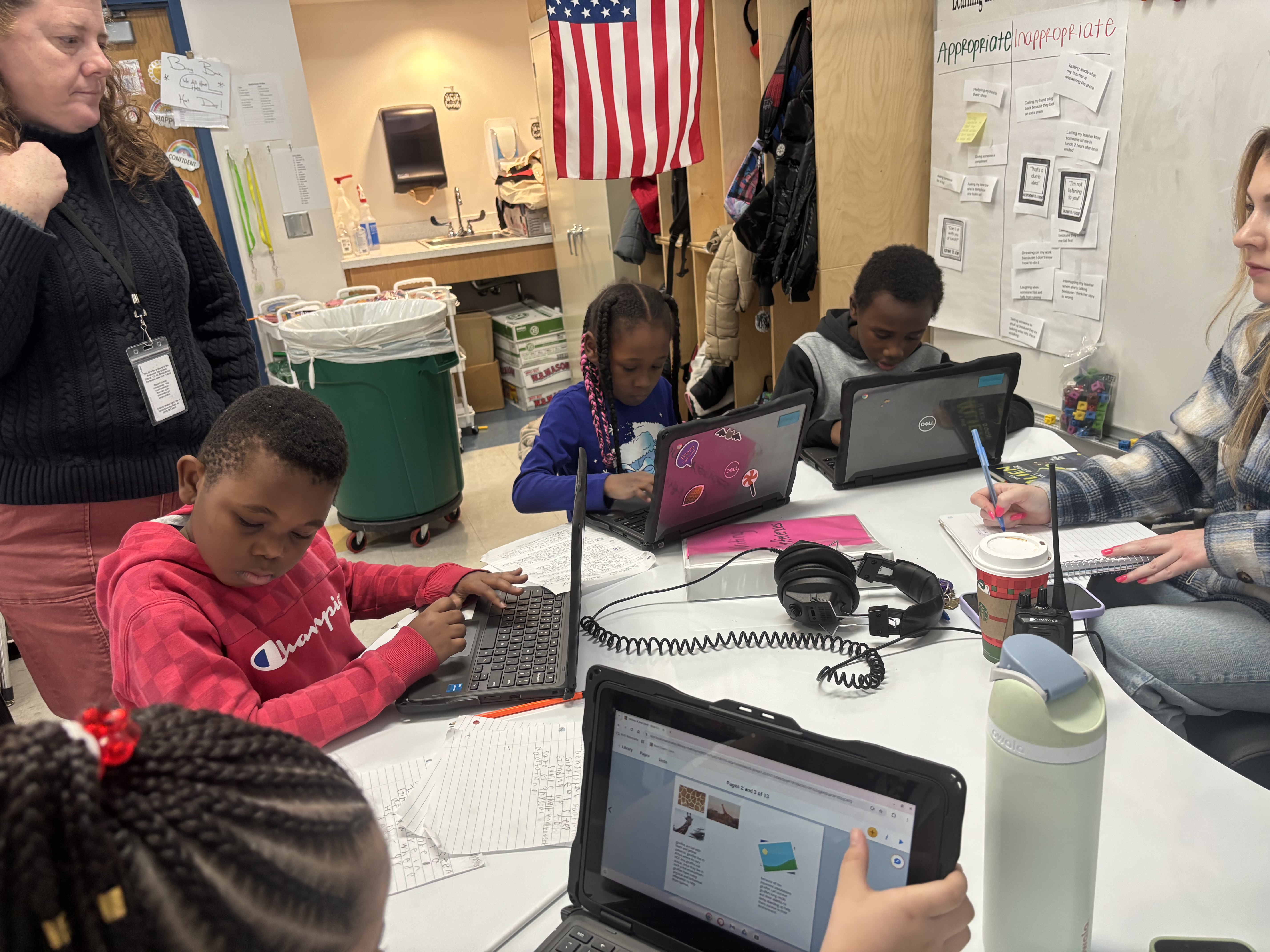 A group of students are working on computers in a classroom setting, with an American flag and various educational materials visible in the background.