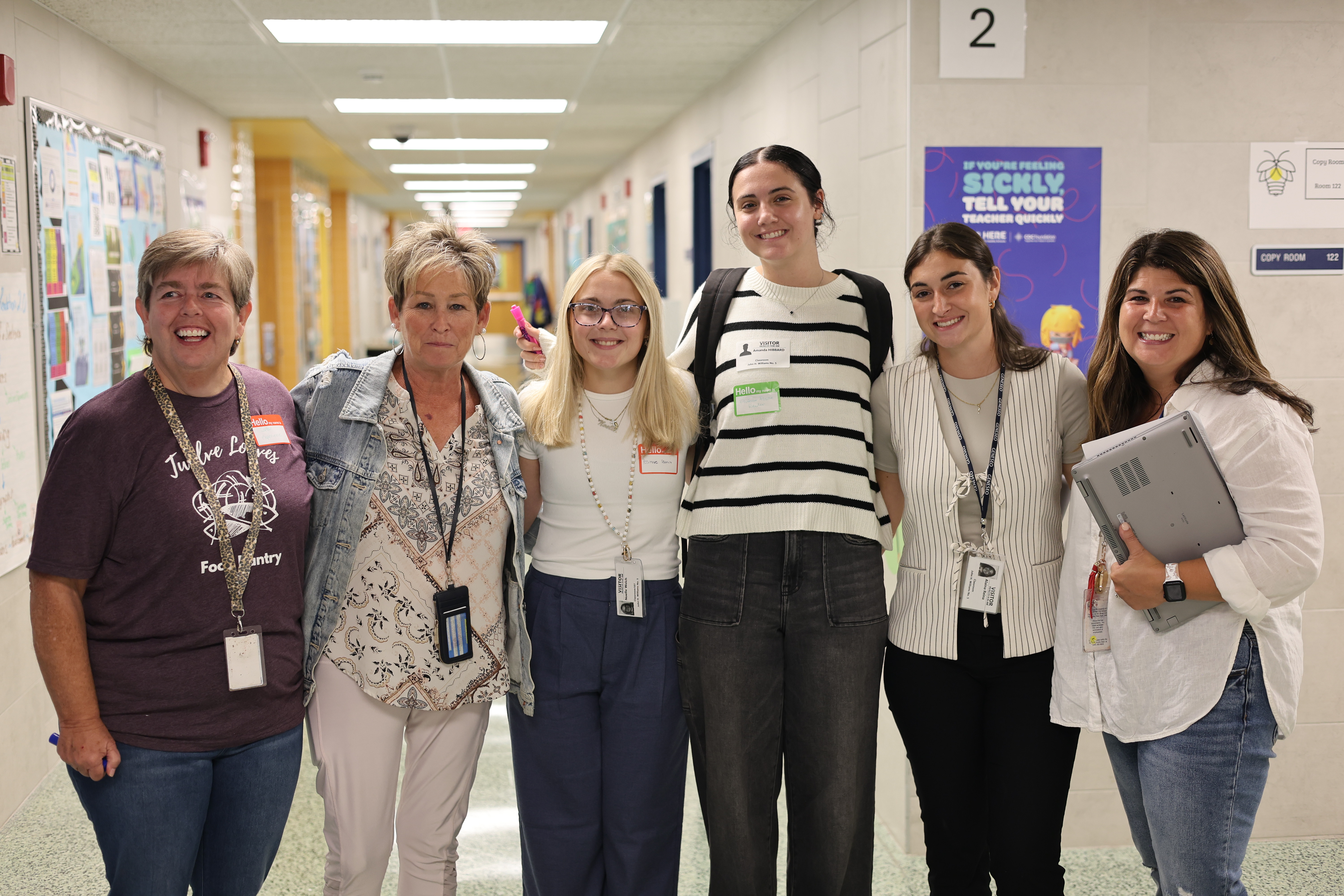 A group of smiling young women standing together in a school hallway, with various posters and signs visible in the background.