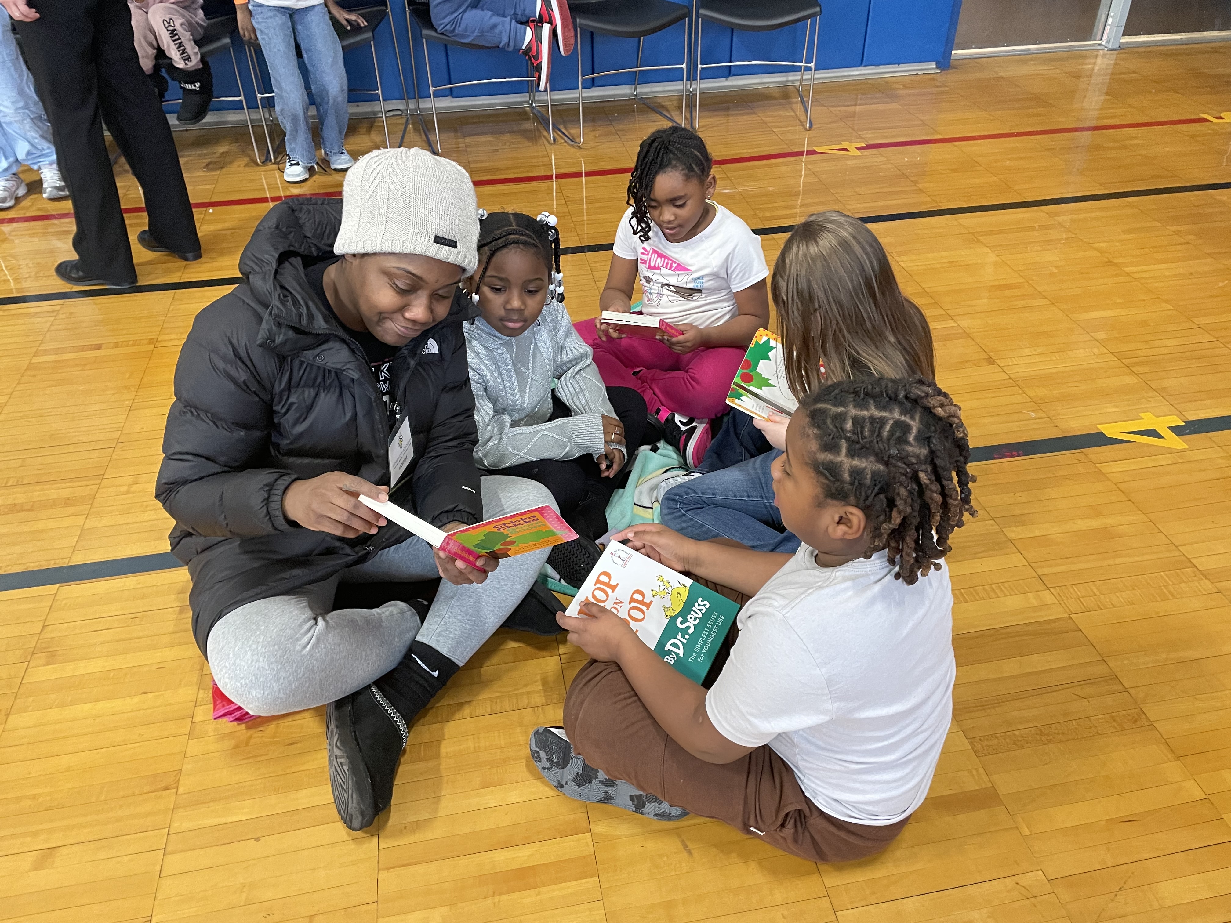 A group of people, including adults and children, are sitting on the floor of what appears to be a school gymnasium, engaged in an activity or discussion.