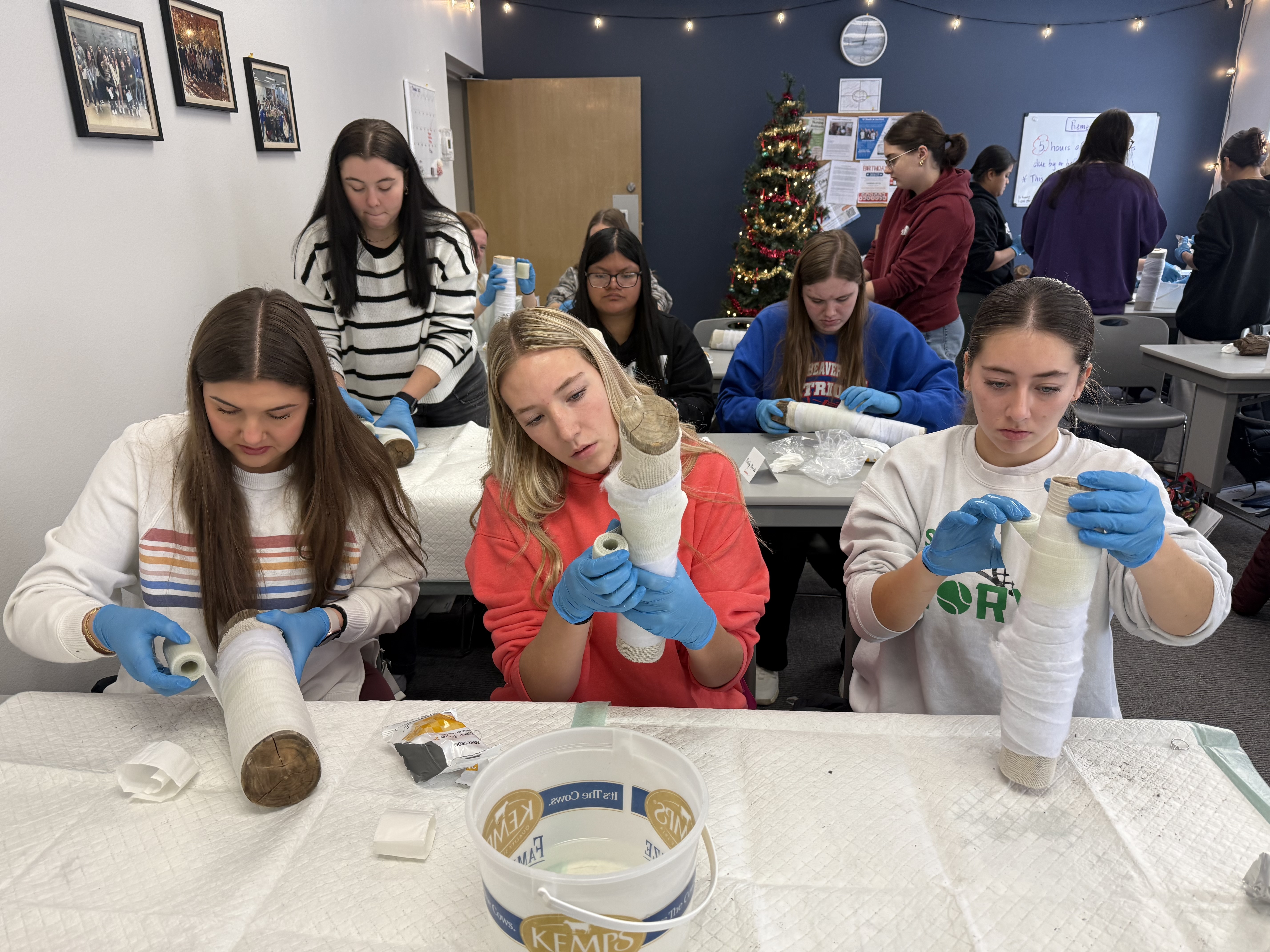 A group of people, some wearing colorful clothing, are gathered around a table, engaged in an activity that appears to involve crafting or creating something. The background includes a Christmas tree and other decorations, suggesting a festive or holiday setting.