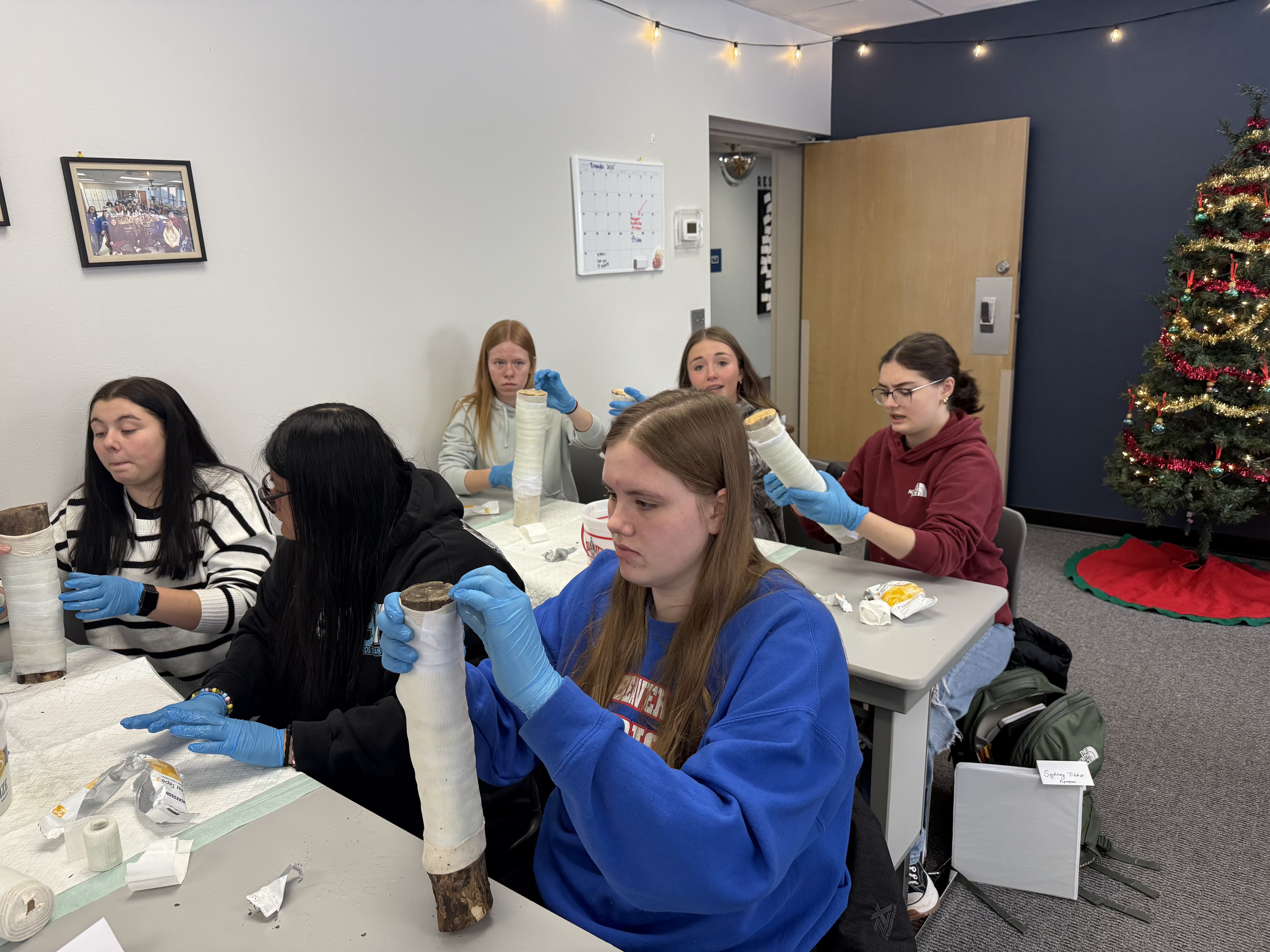 A group of young women wearing blue gloves and aprons are gathered around a table, working on various crafts or activities in a room decorated for the holidays.